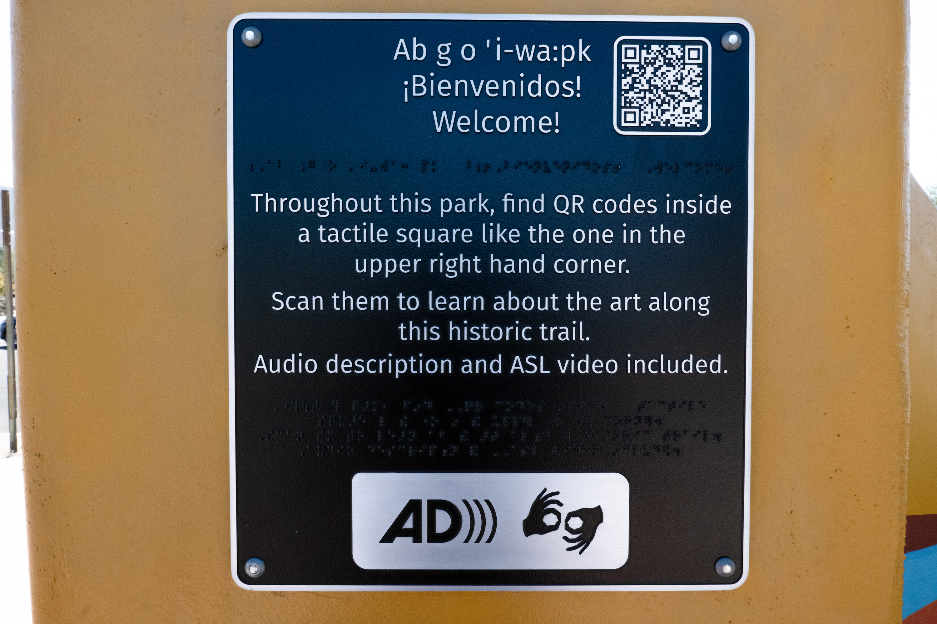 A sign bolted to an orange wall welcomes visitors in three languages and braille. "Throughout this park, find QR codes inside a tactile square like the one in the upper right hand corner. Scan them to learn about the art along this historic trail. Audio description and ASL video included." AD and ASL symbols