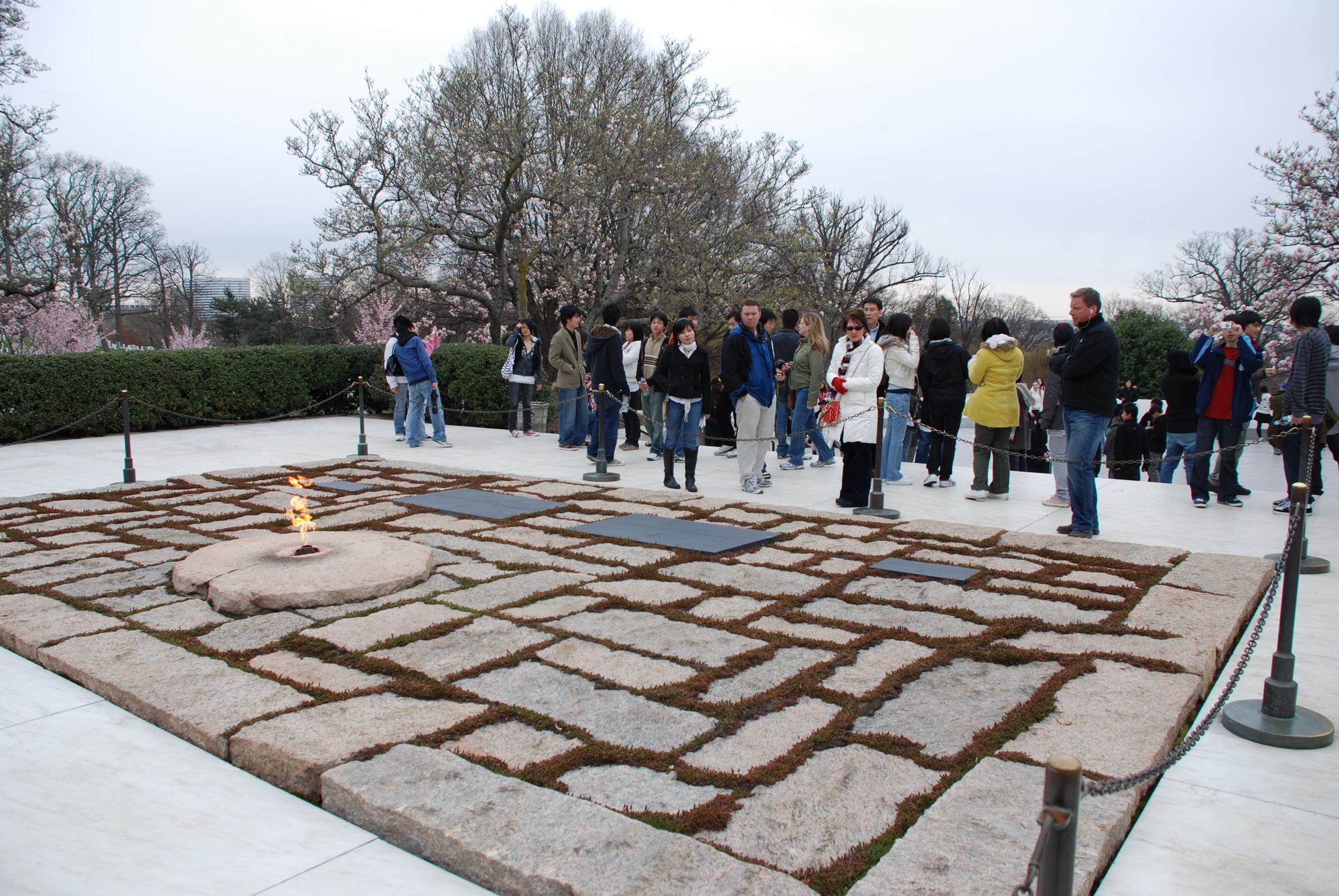A small flame in a patch of stone brickwork is surrounded by ropes. Sever visitors gather around it. 