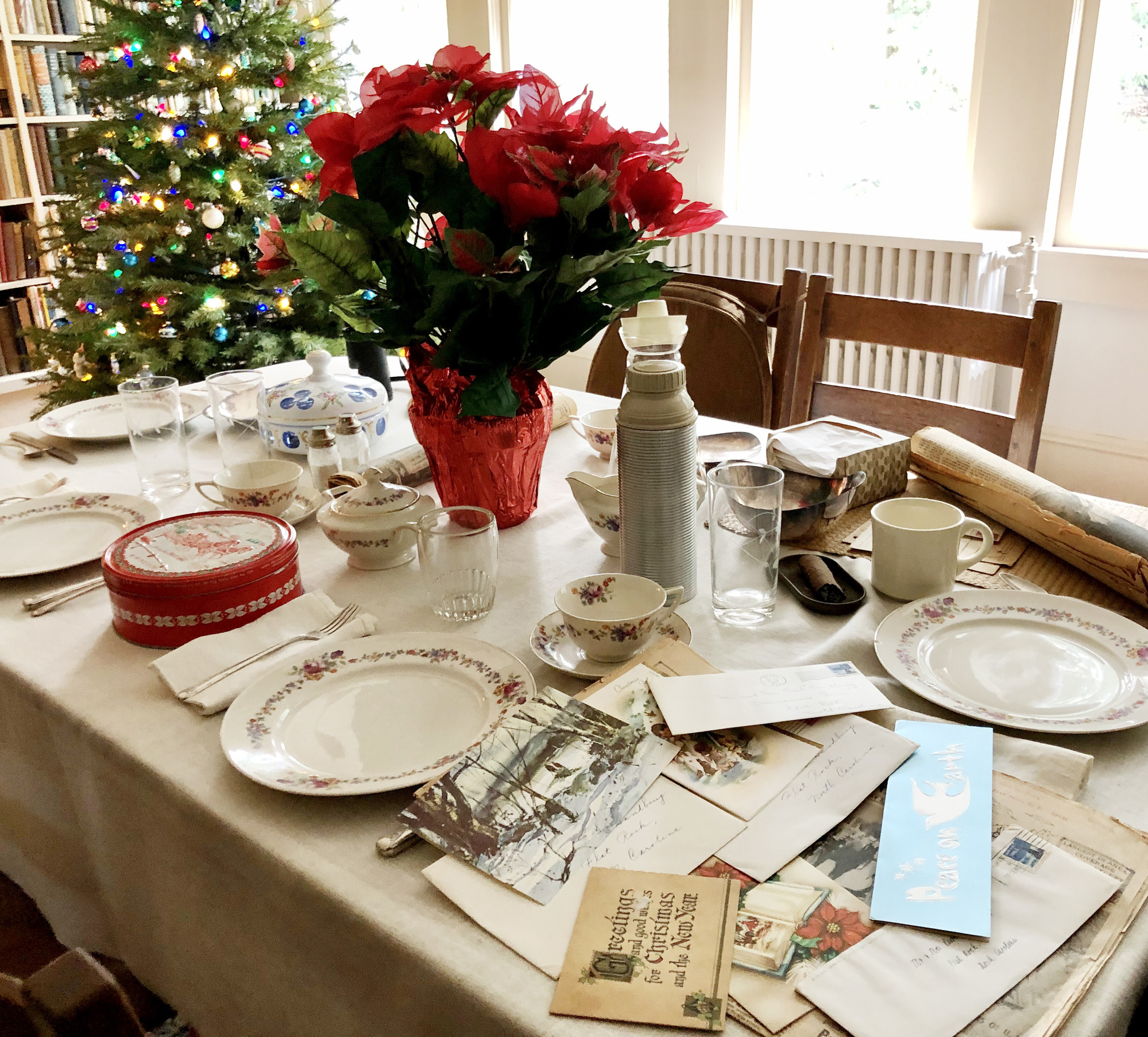 Image of a dining table covered with a cloth and set for dinner. Holiday cards and mail are scattered on a corner, a poinsettia sits in the middle and a decorated Christmas tree is in the background.
