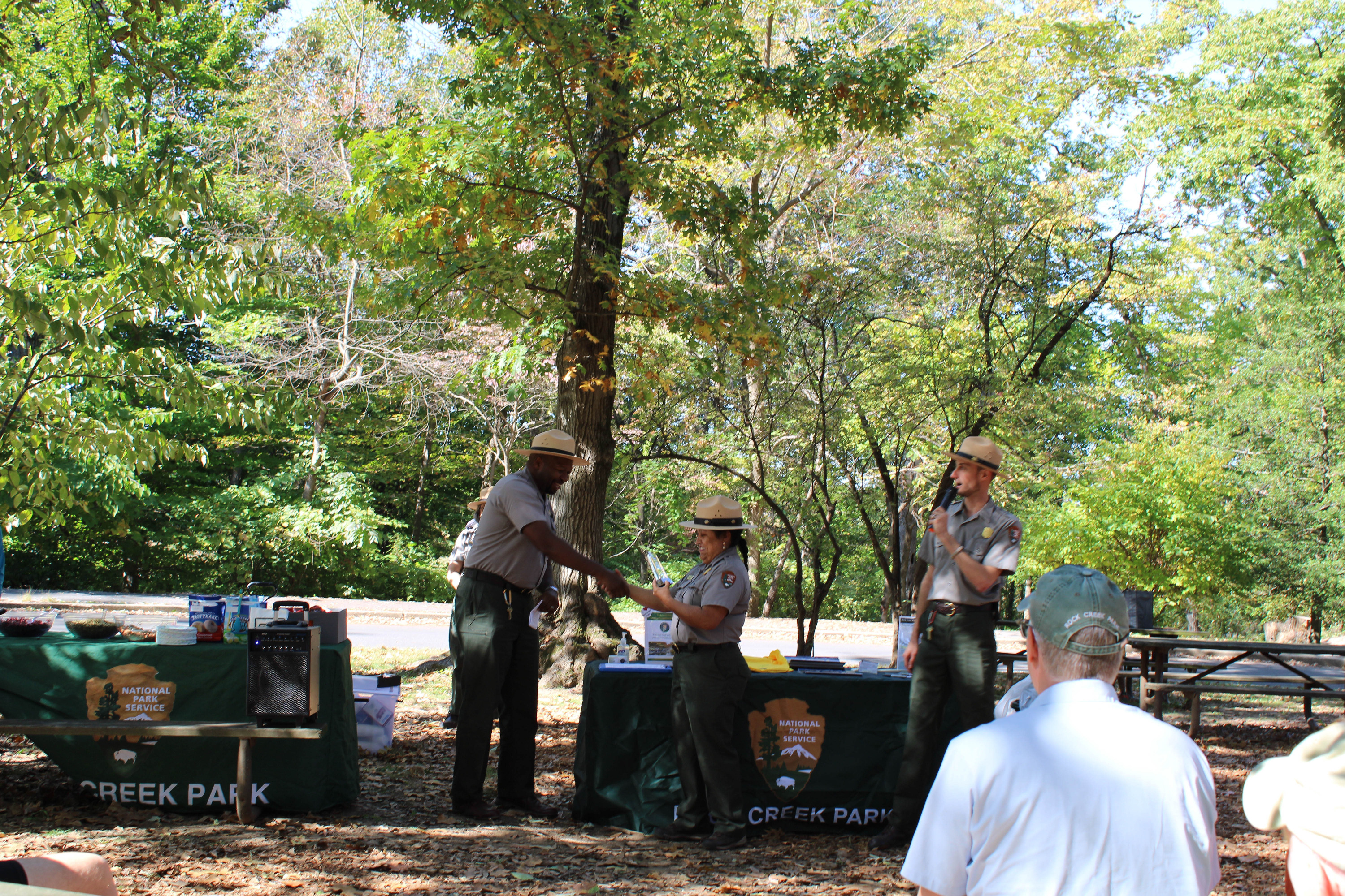 Standing in front of two National Park Service branded tables, a NPS uniformed man shakes a NPS uniformed woman's hand. A crowd of people sitting at picnic tables watch and applaud.