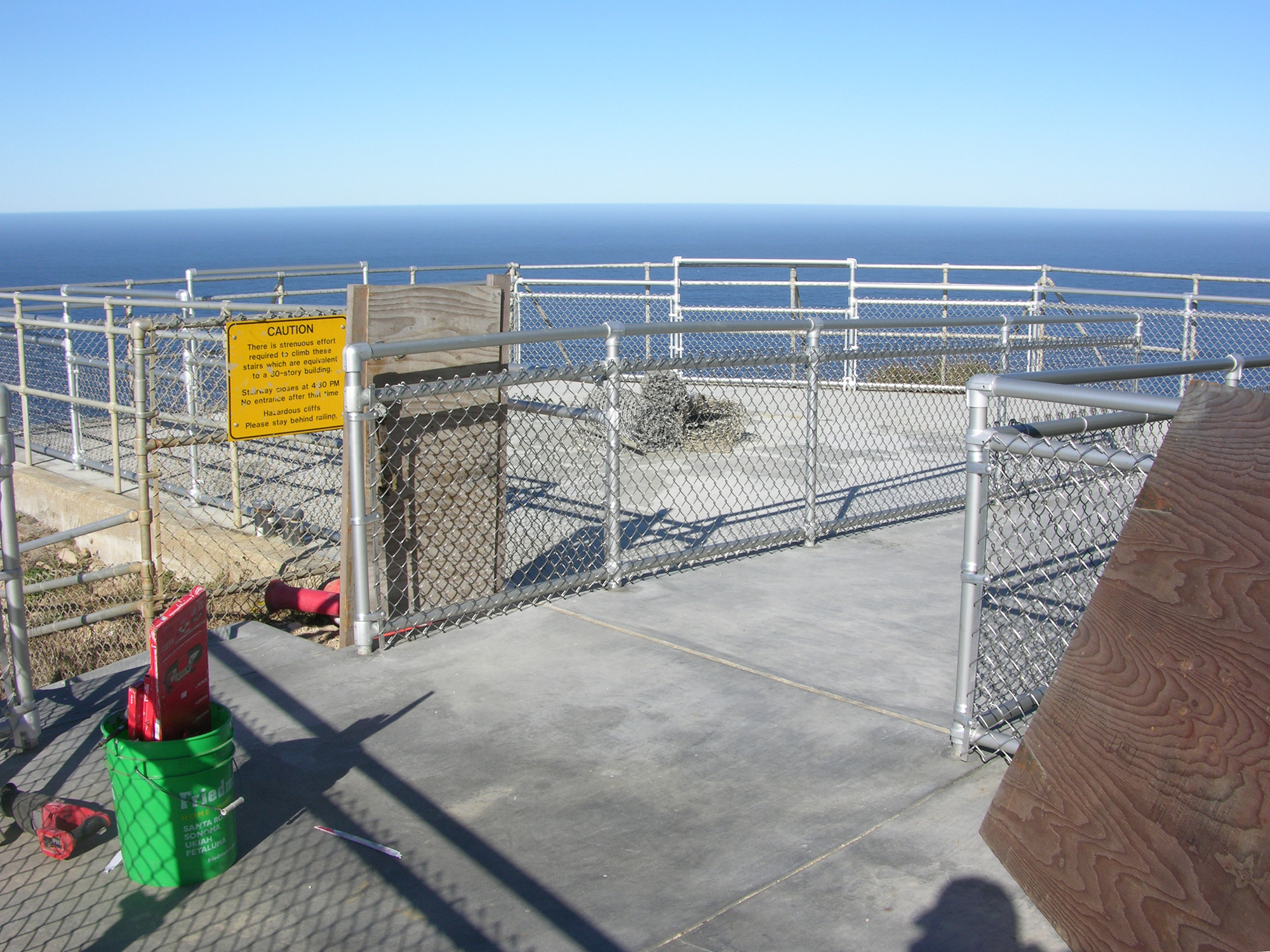 A newly-installed chain-link fence surrounds a rectangular concrete platform. Pieces of plywood, a green bucket and other tools are visible in the foreground. The Pacific Ocean and clear skies are in the background.