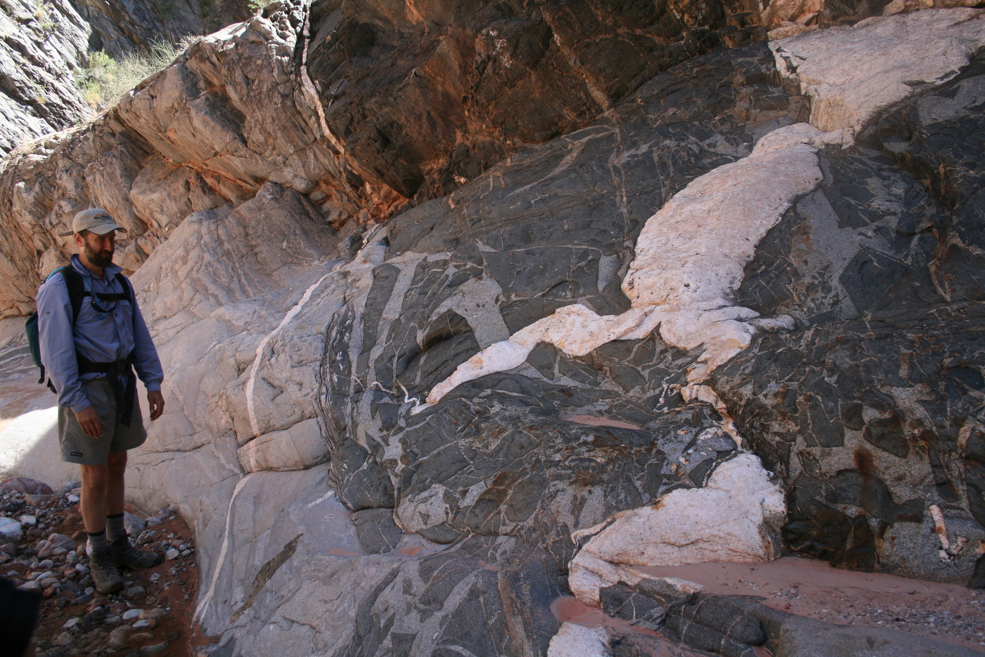 Photo of a person standing by a rock face with light color veins cutting through a mass of darker rock.