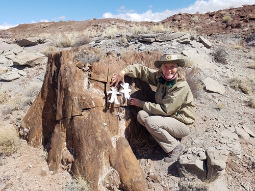 Woman in uniform holding two paper dolls in front of a petrified stump on a hillside under a blue sky.