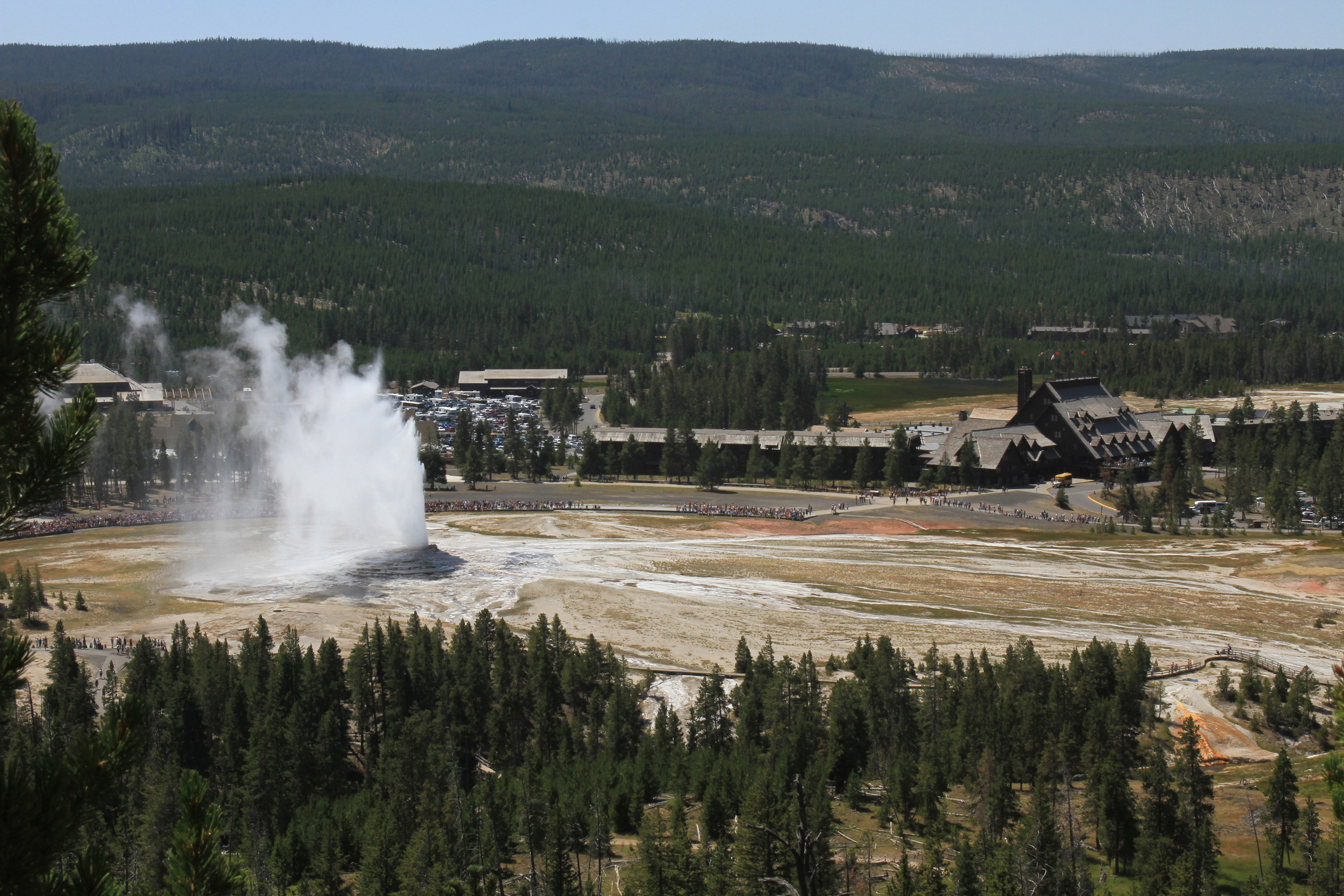 View from Observation Point of visitors watching Old Faithful Geyser with Old Faithful Inn and Old Faithful Visitor Center in background.