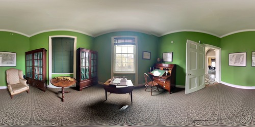 In this 360 image, there is a desk with a chair, two tea tables, one with a portable writing desk and one with five books, a winged armchair, and two bookcases. The walls are lime green.