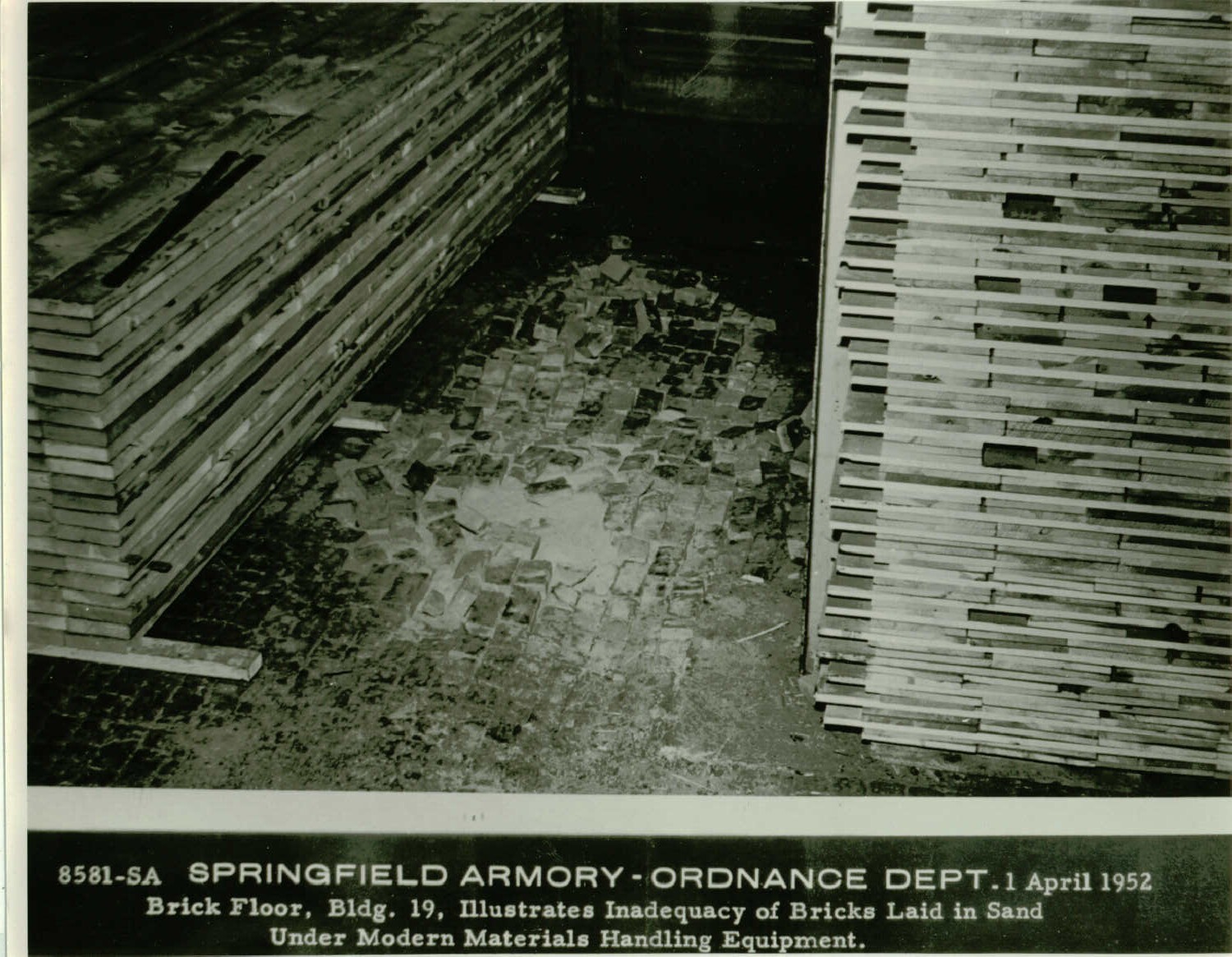Black and white photo looking at a damaged brick floor between two stacks of wooden planks. Text under the photo reads, “8581-SA Springfield Armory – Ordnance Dept. 1 April 1952, Brick Floor, Bldg. 19, Illustrates Inadequacy of Bricks Laid in Sand Under Modern Materials Handling Equipment.”