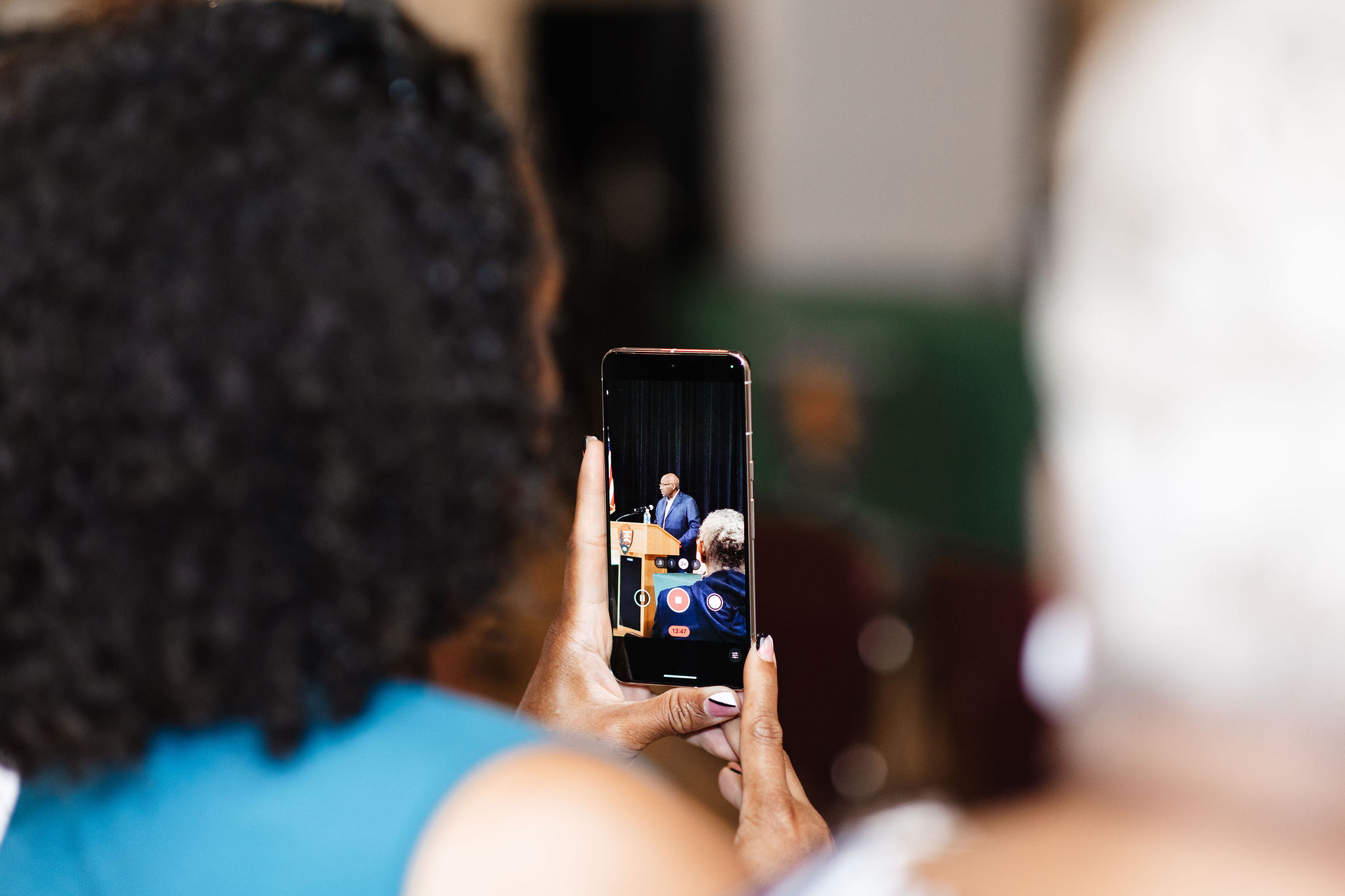 An african american woman in a blue blouse sits with her back to the camera. The photo focuses on a phone in her hands taking a picture of an african american man in a blue suit speaking behind a wooden lectern with the NPS Arrowhead emblem on it
