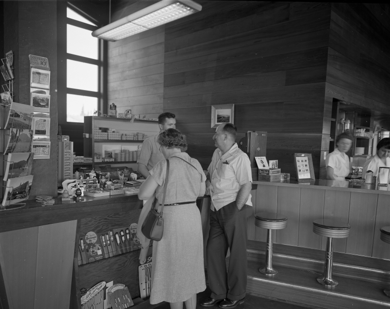 A black and white photo of people at a counter where postcards and souvenirs are on display. A counter with round stools nearby.