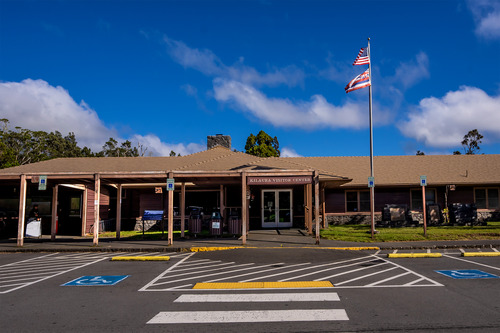 A one-story brown building that serves as the Kīlauea Visitor Center in Hawaiʻi Volcanoes National Park. The American flag, with the Hawaiʻi state flag below it, flies on a flagpole in front of the building. 