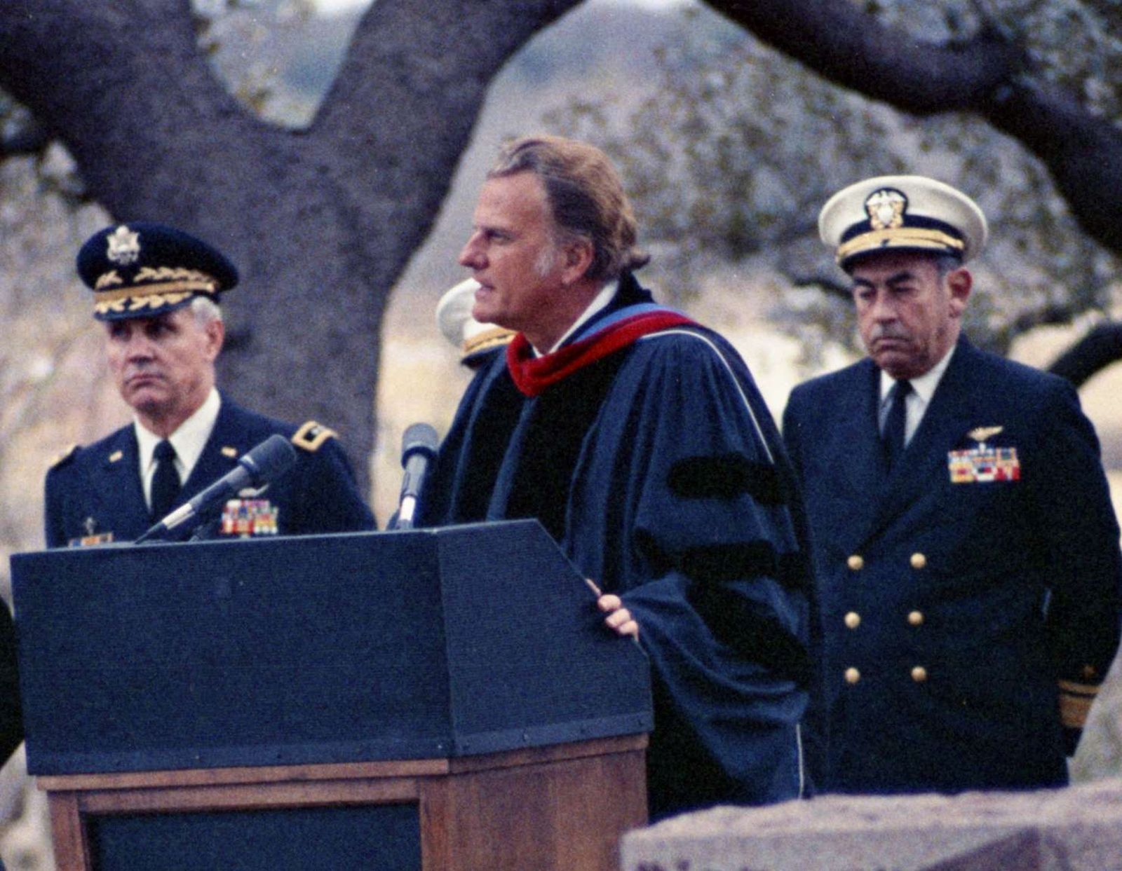 An older man wearing dark blue clerical robes stands at a lectern with microphones. Behind him are two officers in military uniform.