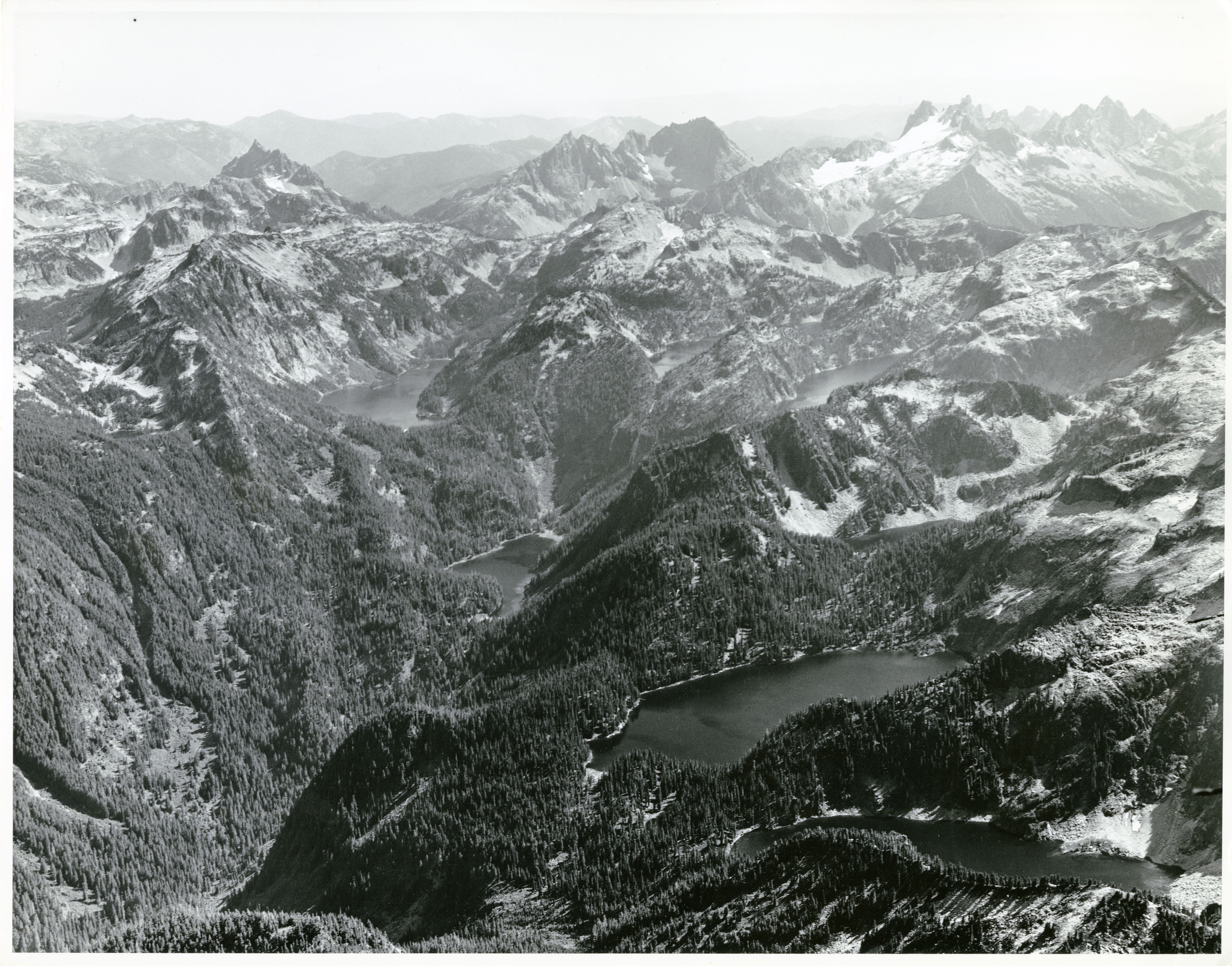 Aerial view of forested mountain peak. A variety of forested and snow covered ridges, several lakes between ridges.