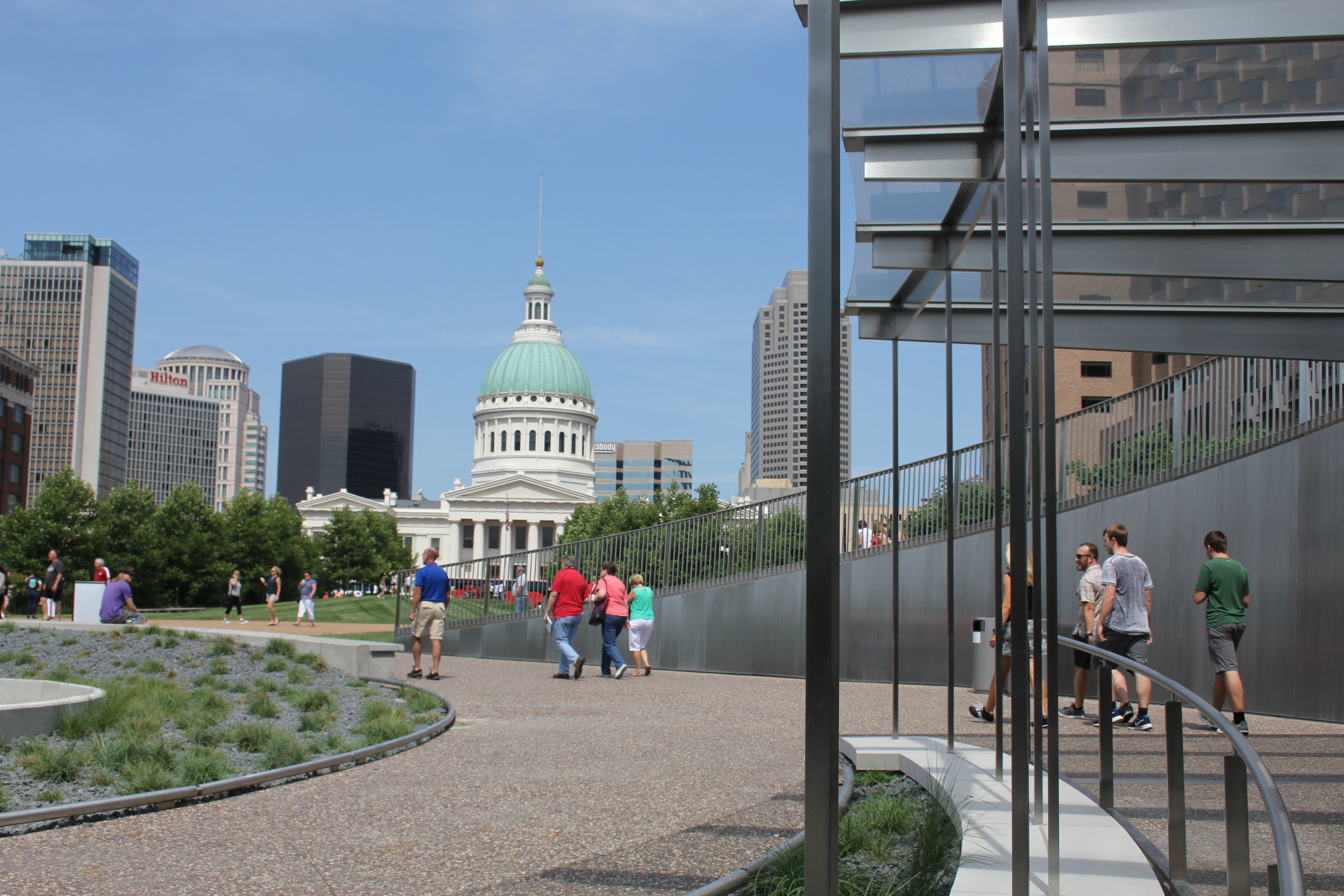 Visitors walk out of the Arch visitor center along a curved sidewalk. The Old courthouse sits in the background.
