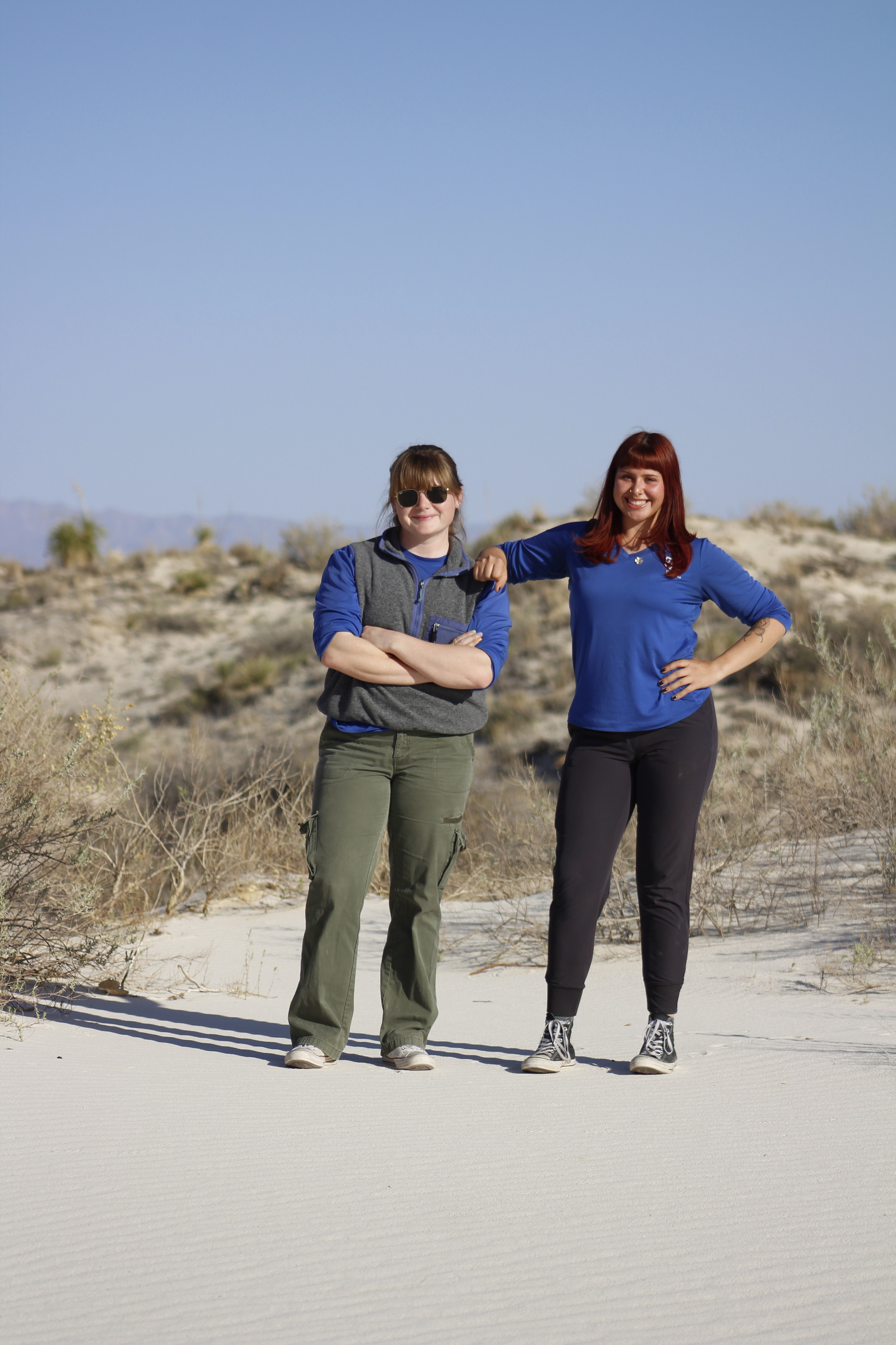 Two women pose side by side on vegetated white sand dunes under a clear blue sky. 