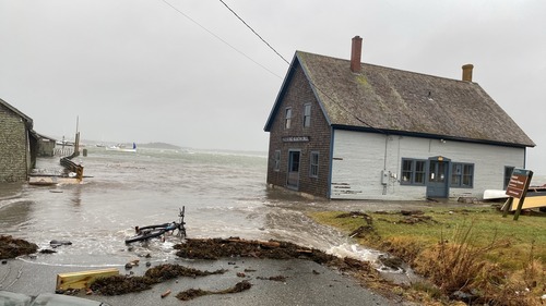 A small wooden building covered in shingles is affected by storm tides. The water completely encroaches on the base, and reaches up the road. 