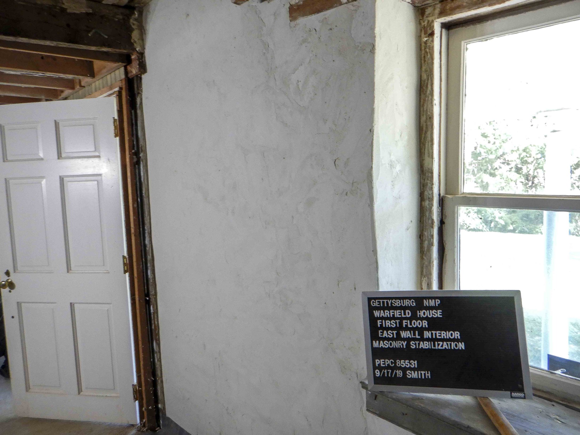 An interior stone wall is cover by a final coat of white plaster. There is a white door on the left and a window on the right.