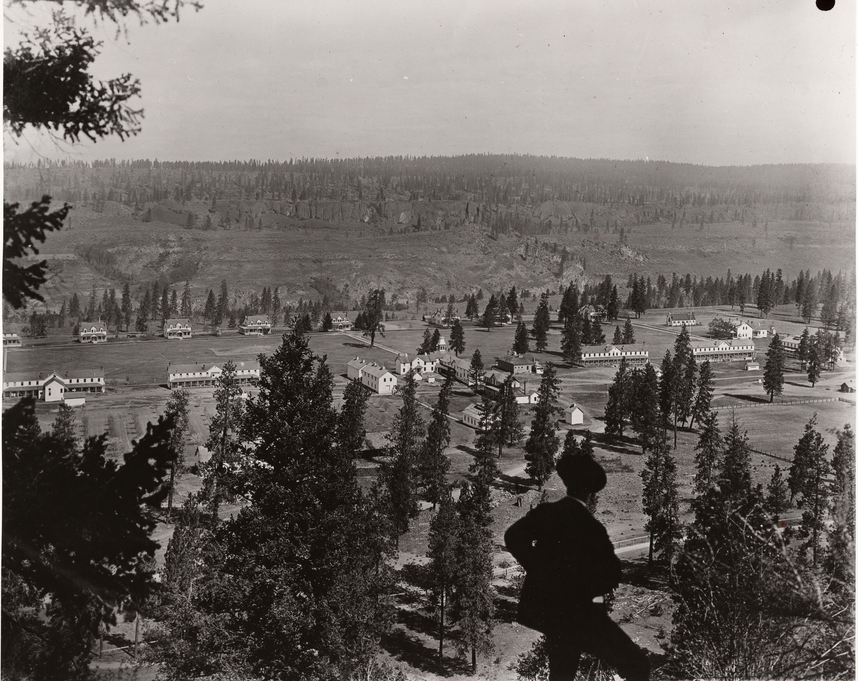 Black and white photograph of a man looking over a complex of buildings in a valley below