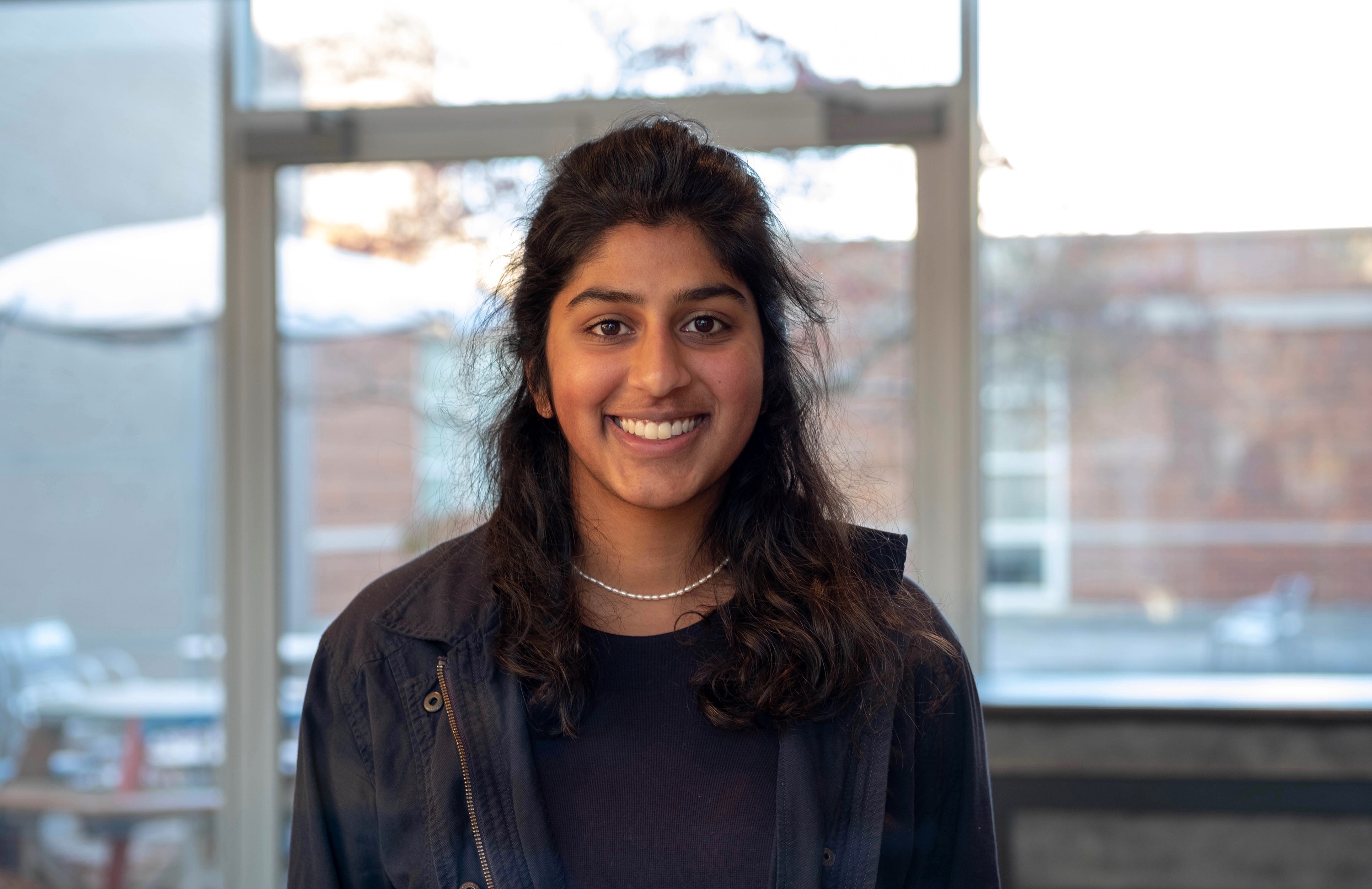 A young woman with curly, dark hair and black jacket stands smiling in front of a window.