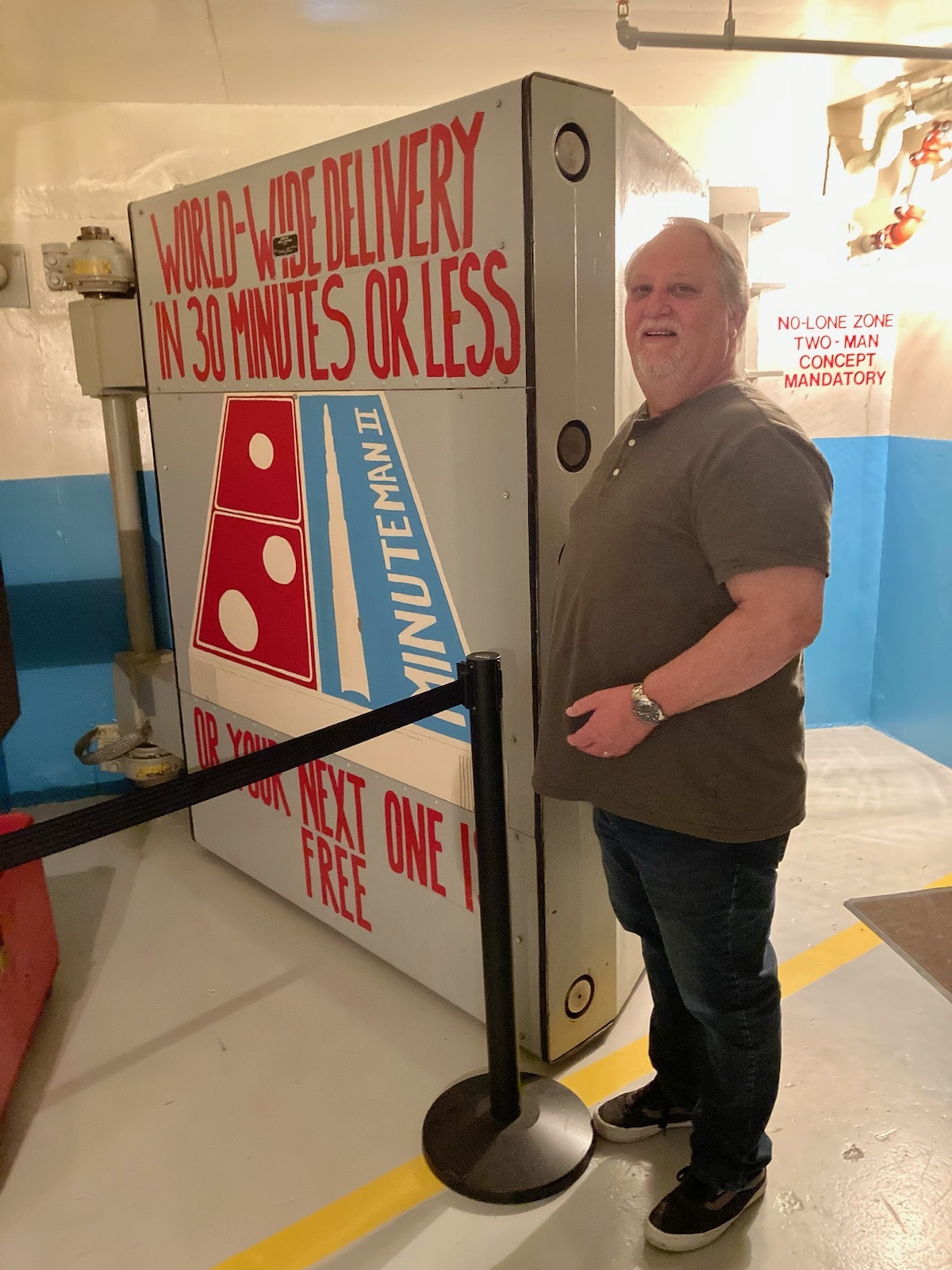 
Man in a gray shirt and blue jeans standing next to a large concrete door with a red white and blue domino's pizza logo on it and the words in red: World wide delivery in 30 minutes or less or your next one is free. 
