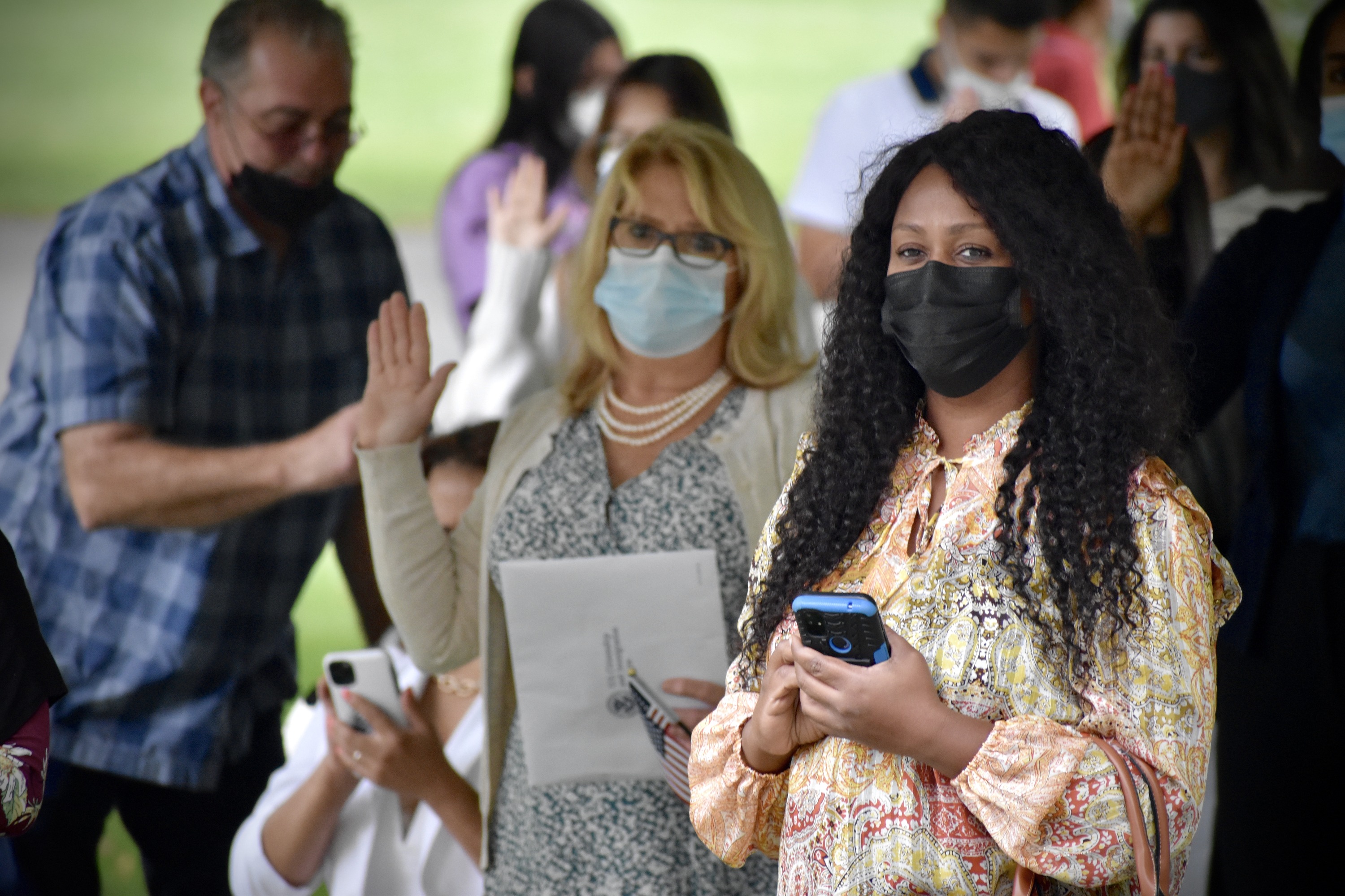 Individuals stand underneath the tent with their right hand raised taking an oath. A woman is in the foreground with her phone in her hands. 