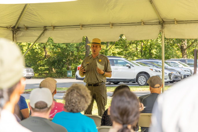 A tall man in a park ranger uniform gives a speech in front of a crowd that sits under a tent. 