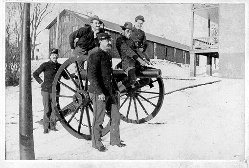 The cannon and soldiers are near several buildings which are in the background.  Two of the soliders are standing, one at the back of the cannon and one in front.  The other three men are "on" the cannon.  On is standing on the trail of carriage, one is sitting on the wheel, and the third man is sitting on the barrel.