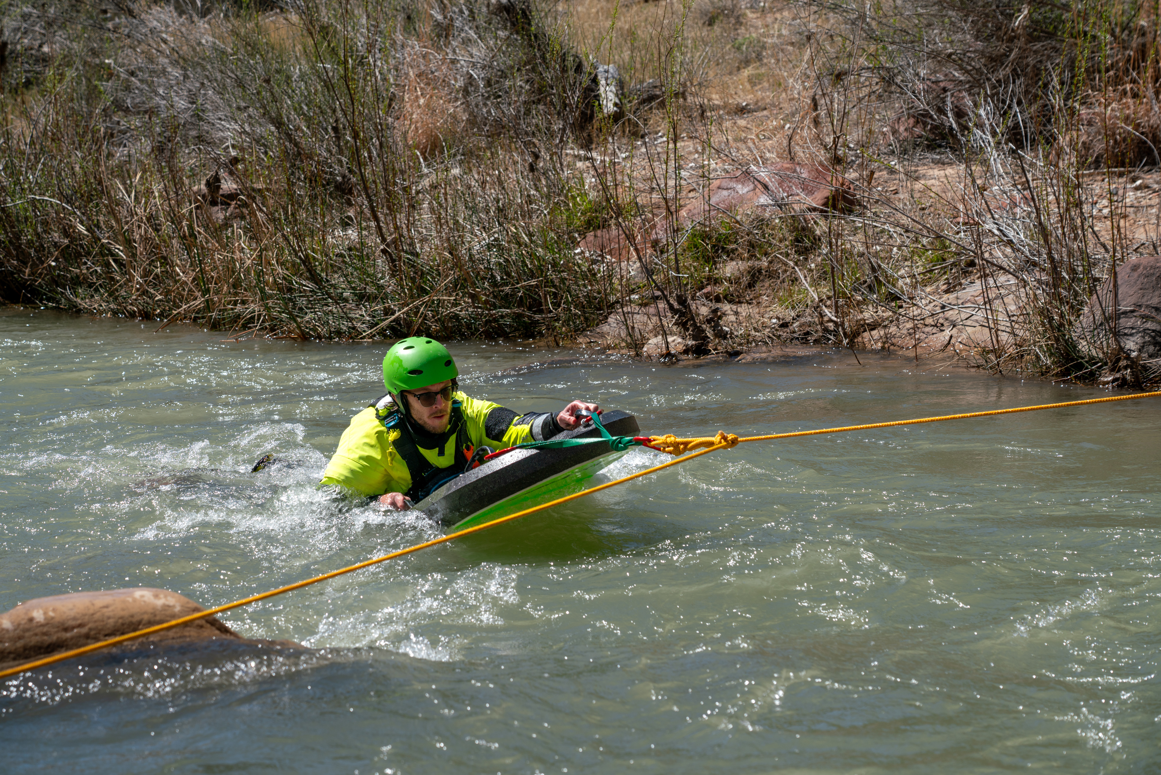 A SAR team member on a boogie board with ropes attached to it in the river. He wears a lifejacket, yellow dry suit, and helmet, with sandstone rocks behind him.
