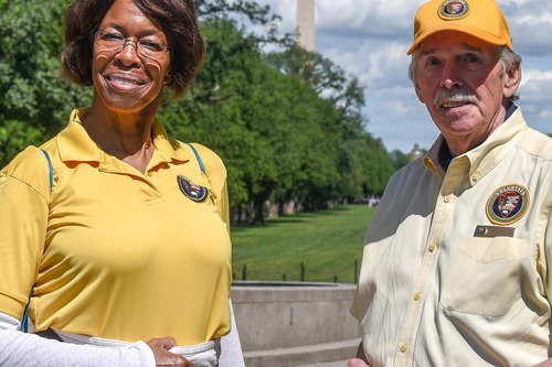 An African American woman with brown hair and yellow glasses is wearing a yellow polo shirt while posing for a photo.

An white man with gray hair is wearing a yellow hat and light yellow button up shirt poses for a photo. Both shirts have NPS volunteer program patches on them. In the background the Washington monument can be seen. 