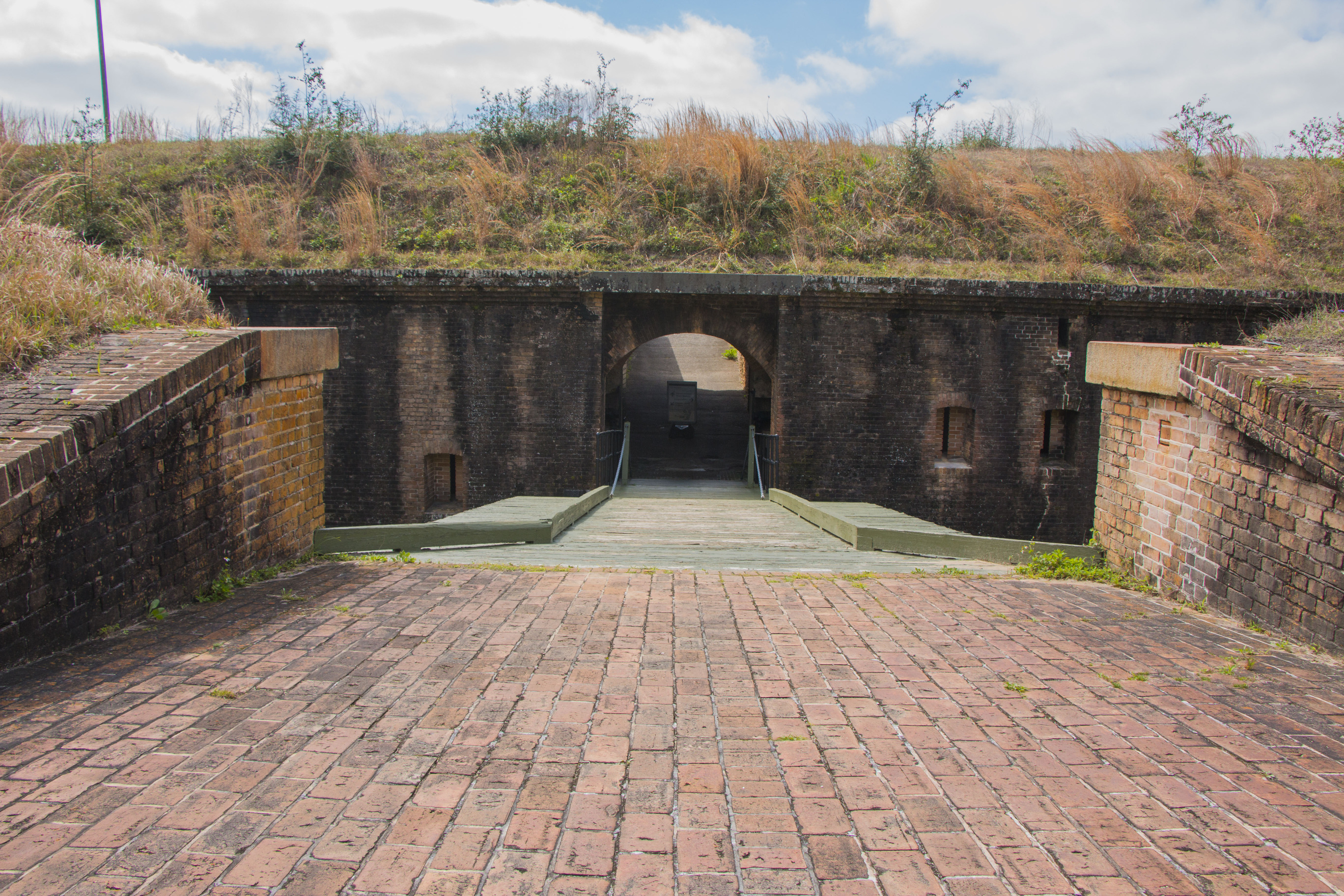 A wooden draw bridge extends across a moat into a wide opening into a masonry fort.