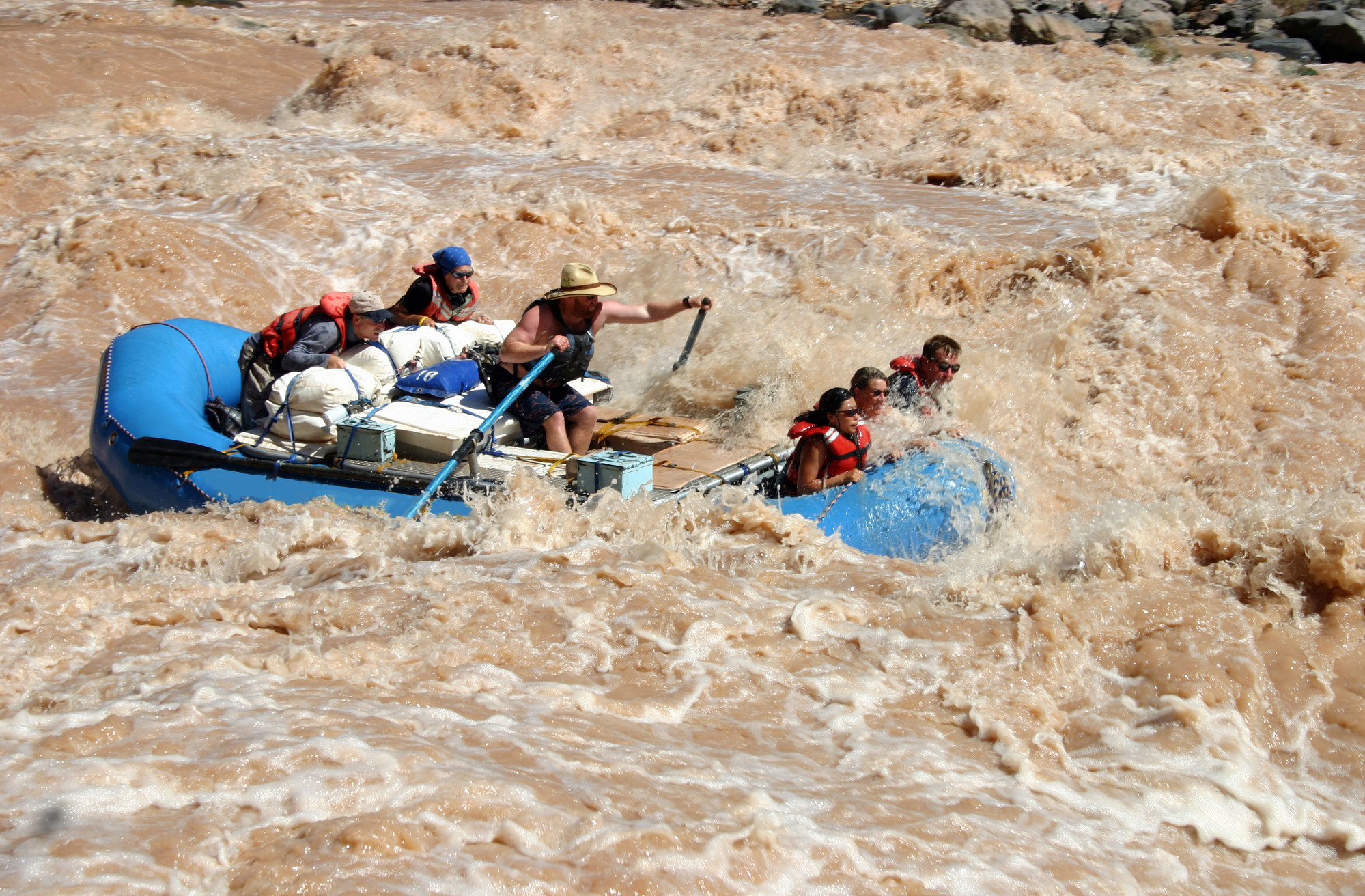 Photograph of six people in a blue raft running through the muddy brown water of Lava Falls Rapid.