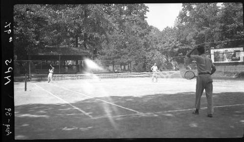 Black & white nitrate negative, interpositive, safe film negative; Whittington Park. Two men playing tennis; tennis pavilion in left midground; telephone poles & large Coca Cola billboard in right midground; cars in center, parked along iron fence & gates.