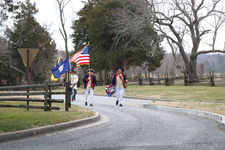 Four men dressed in colonial era clothing, three holding flags. Man in front of the line holding a red, white, and blue wreath.