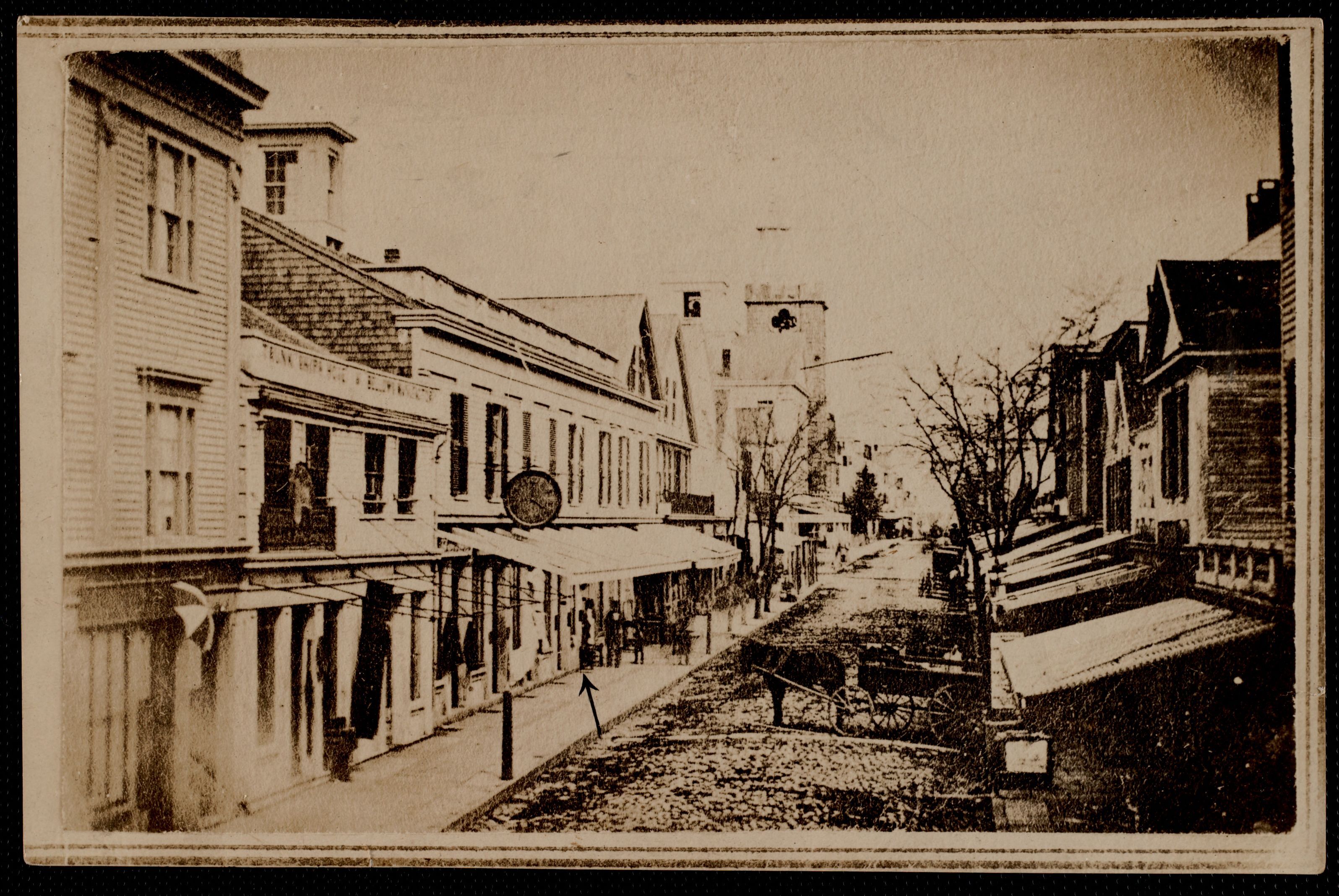 Black and white photograph of a street scene in New Bedford, Massachusetts. A cobblestone street runs through the middle with buildings on either side and pedestrians and horse drawn carriages on the sidewalk and street.