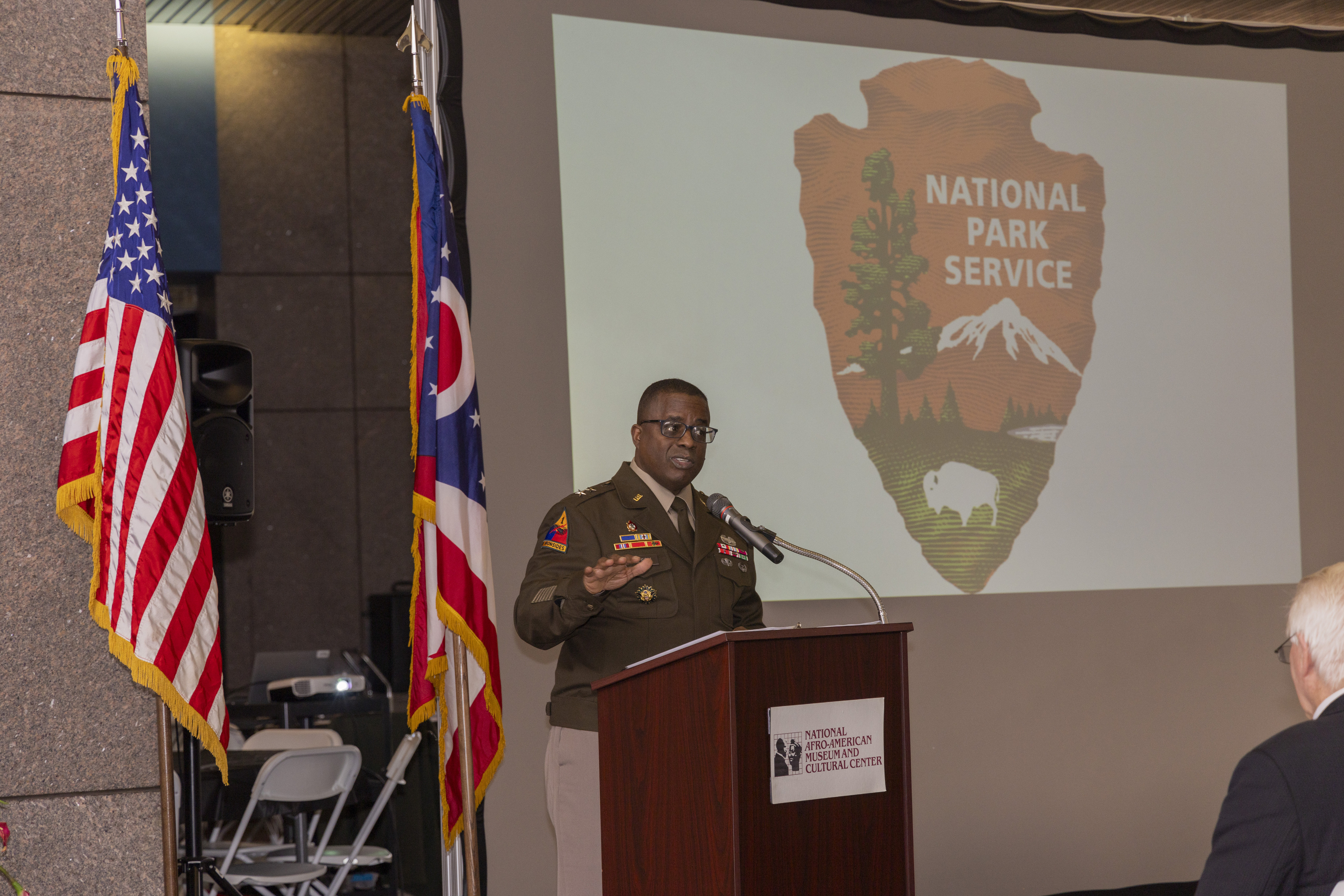 A man in a dark brown uniform stands at a podium and speaks into a microphone.