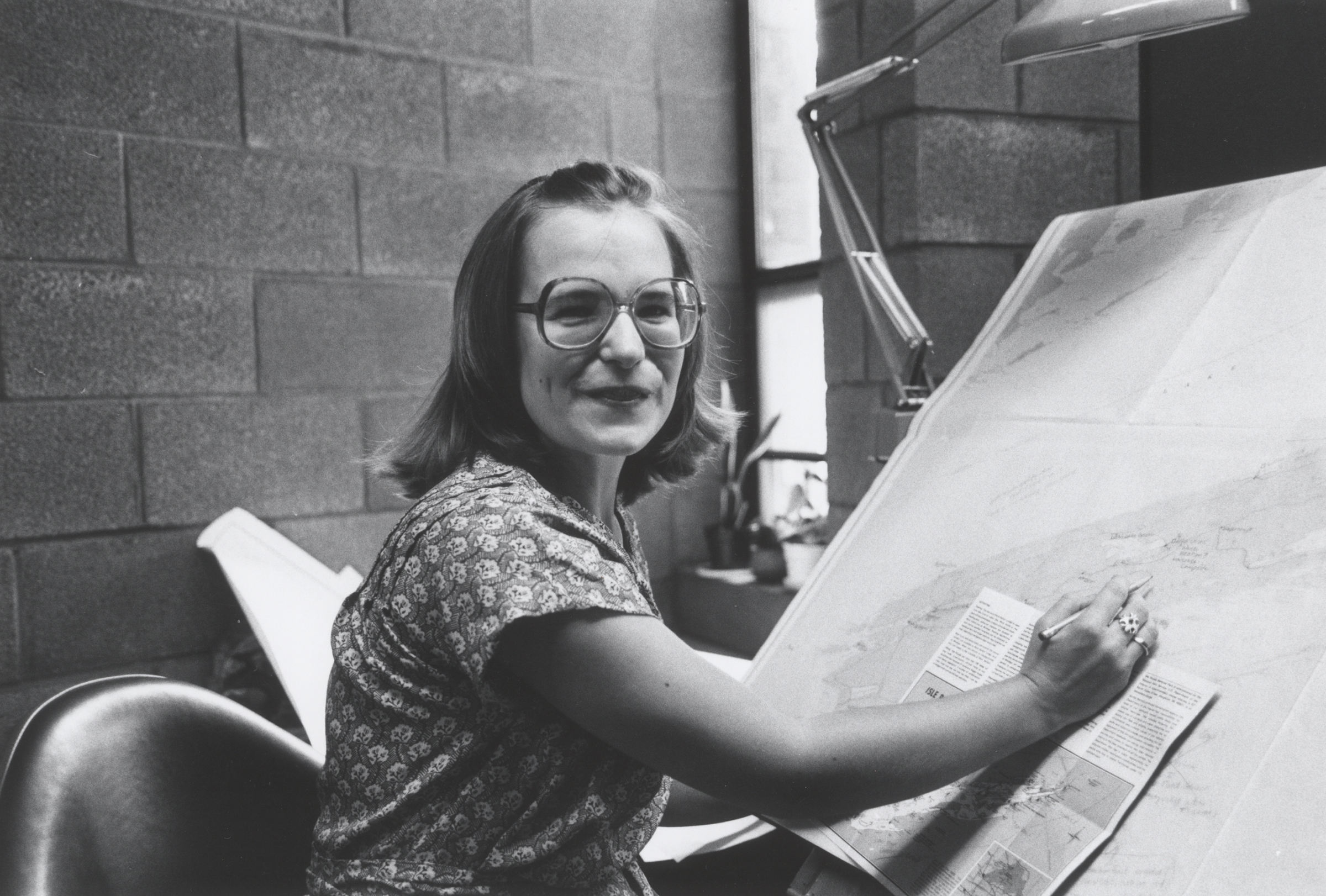 Nancy Haack sits in a chair at a slanted table with a pencil in her hand as she points to a map in front of her.