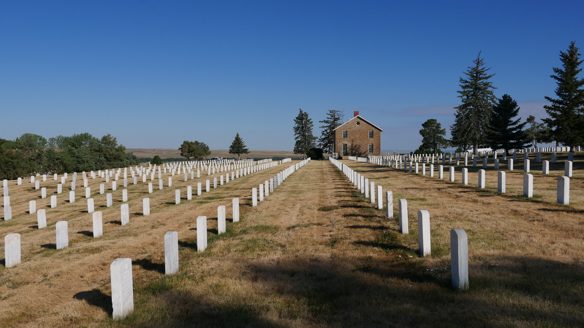 Dormant golden turf grows between evenly spaced rows of grave markers. A two-story stone structure is in the background.