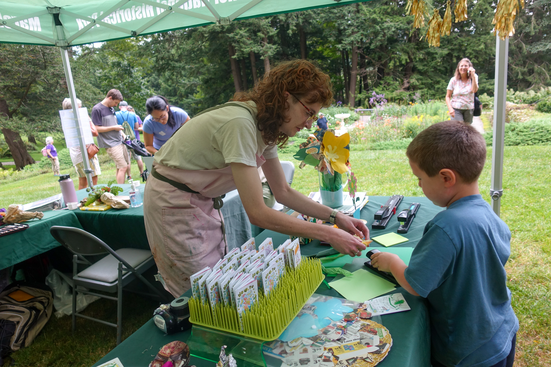 Park intern in pink apron helps a young visitor staple an art project