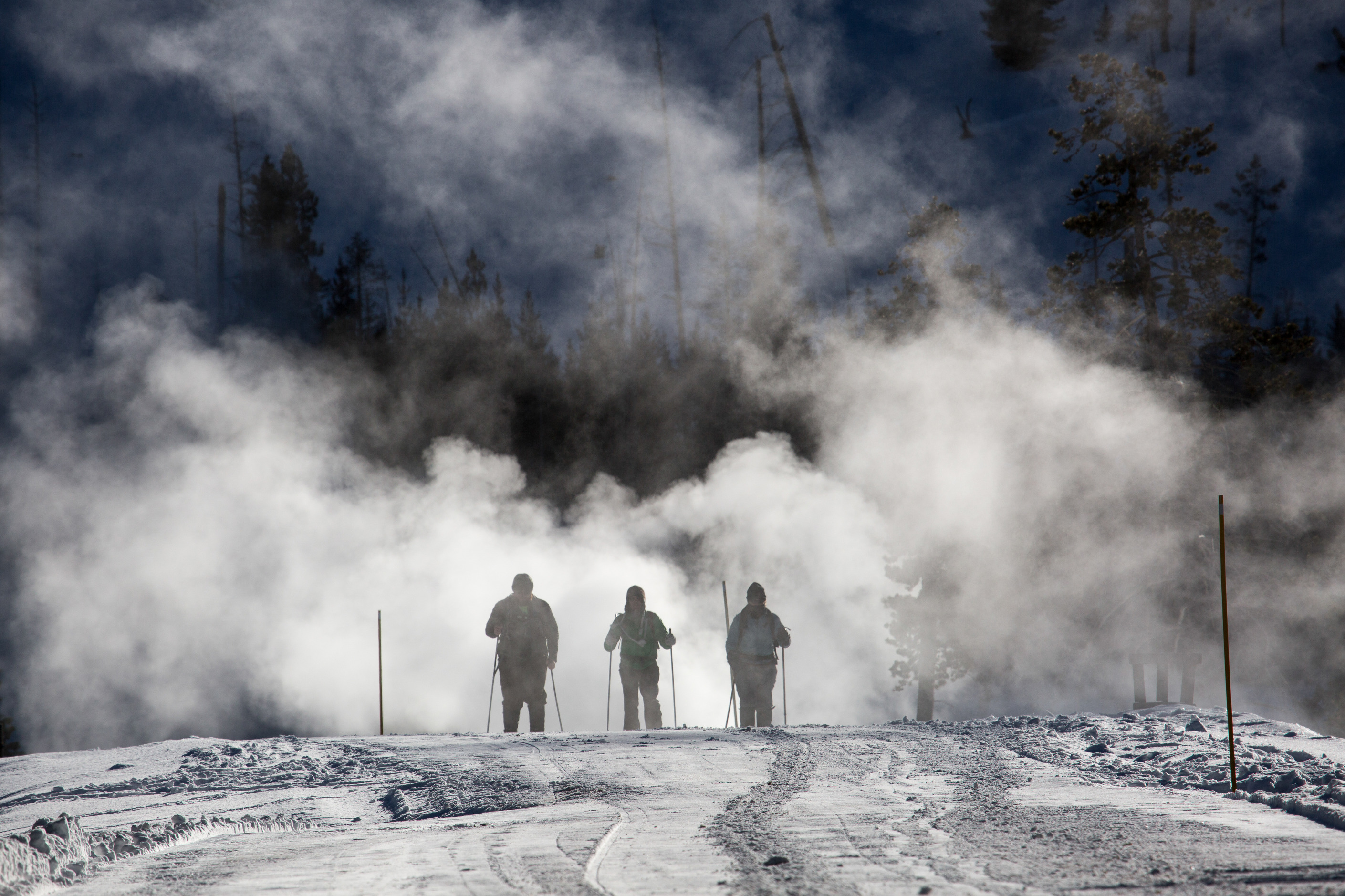 Three skiers are skiing towards camers on a groomed road with steam in the background