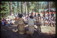 Two women stand at a table with Atsugewi items in front of a crowd of people.