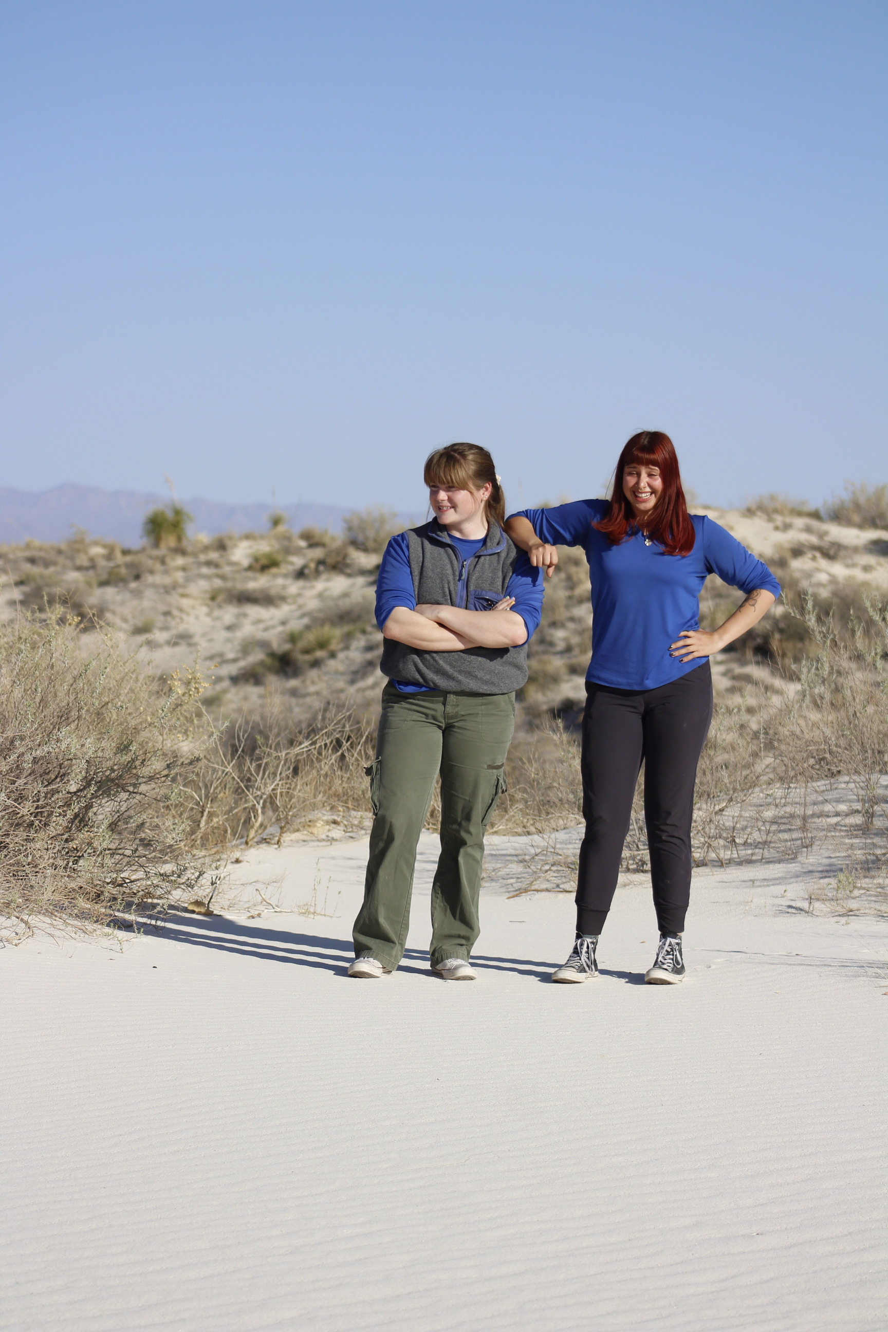 Two women pose side by side on vegetated white sand dunes under a clear blue sky. 