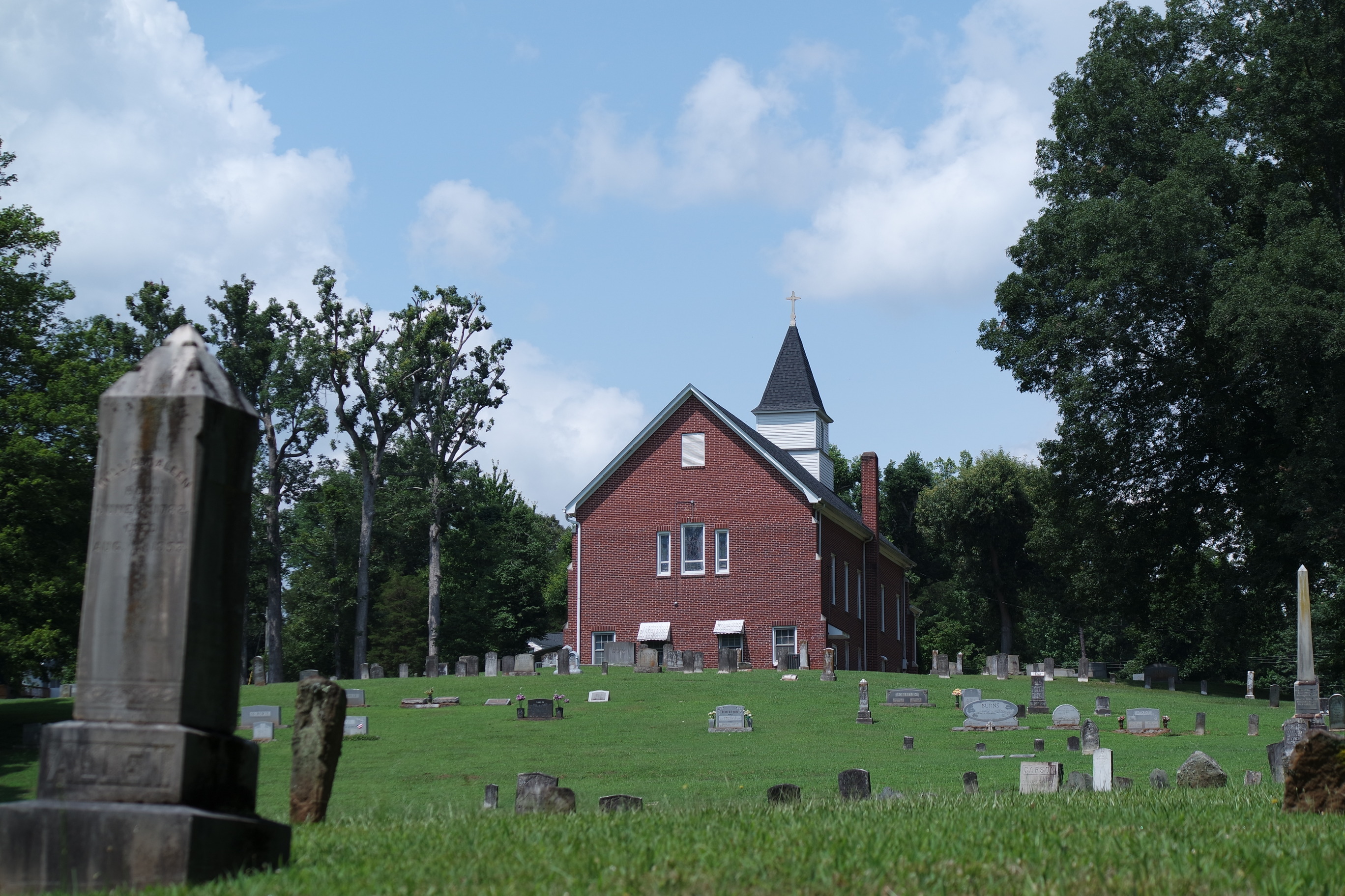 A green grassy cemetery dotted with grey headstones. A brick church is in the background with a white steeple and grey roof. 