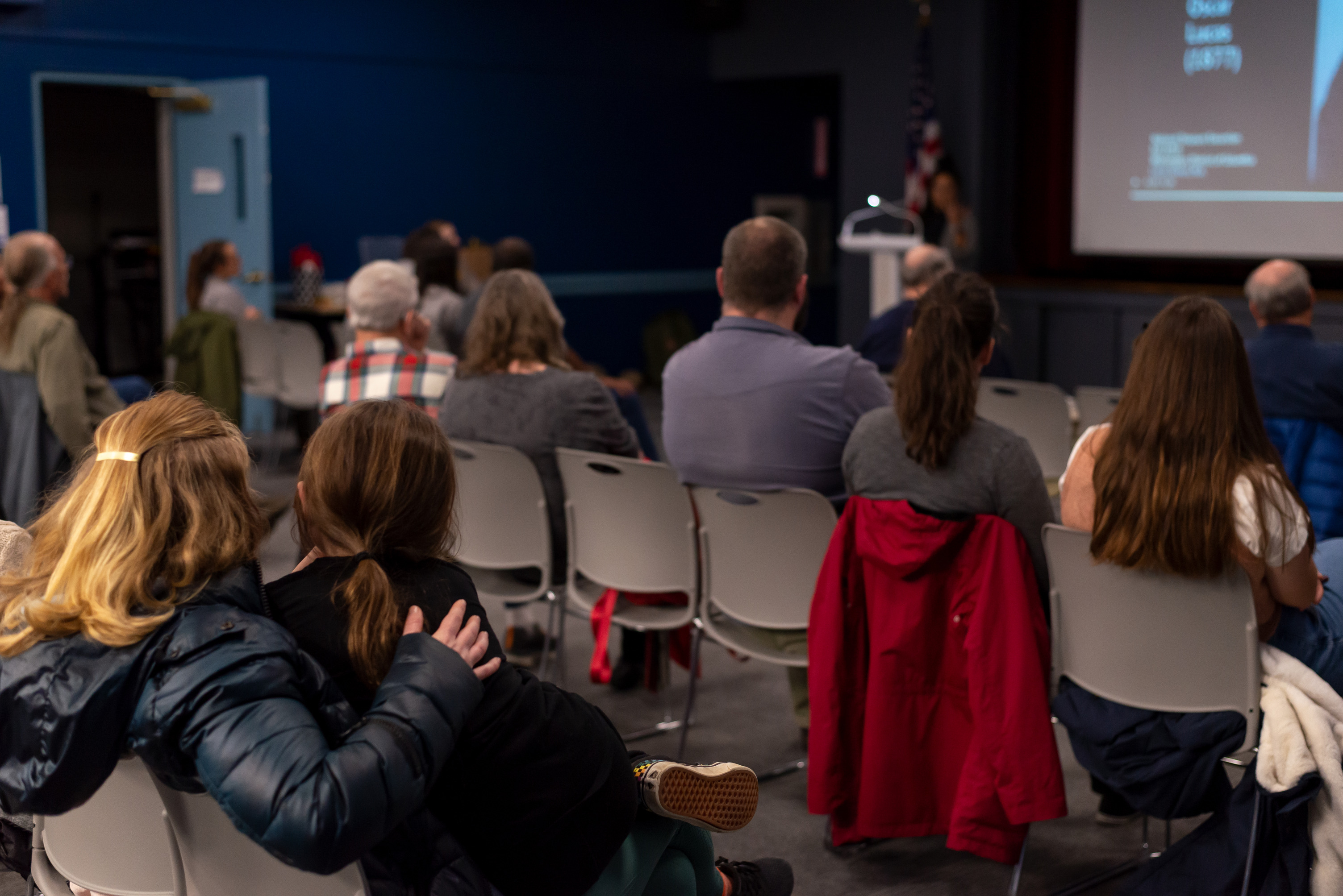 A mother and daughter listen to a speaker lecture indoors.