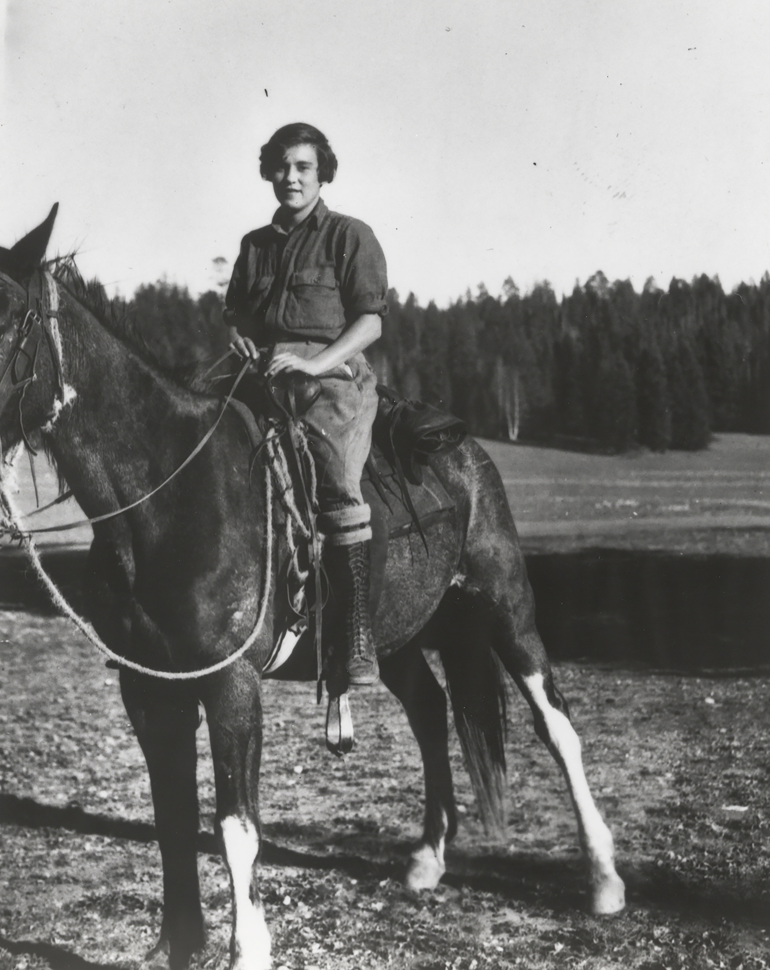 Pauline Mead sitting astride a horse in a field, wearing a button-up shirt with the sleeves rolled up, riding pants and boots. 