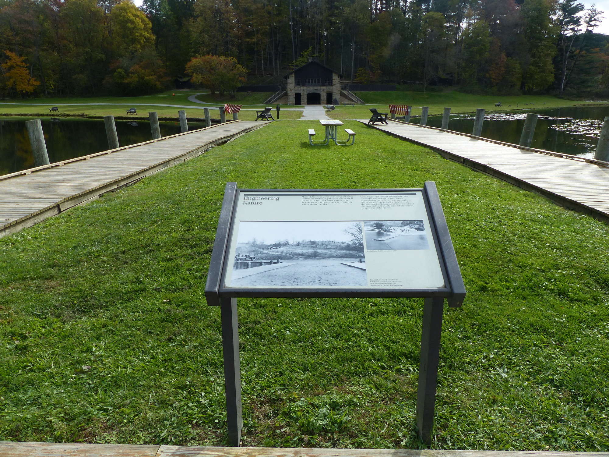 Informational panel on a wide grass pier edged by wooden boardwalk; rustic stone shelter in the distance.