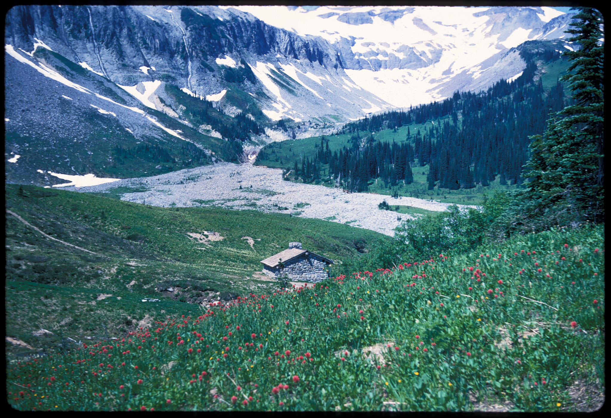 A small stone trail shelter in a broad subalpine meadow in a valley. 