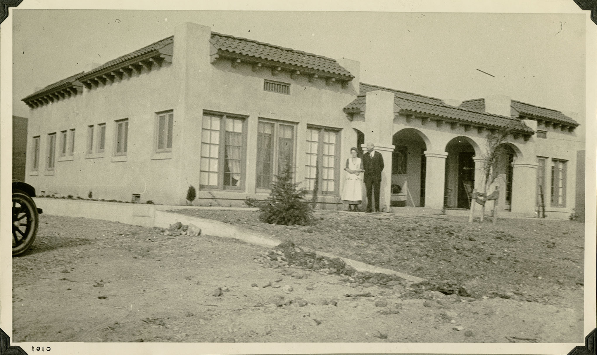 This is an historic black and white photograph from the Scotty's Castle Historic Photograph Collection, Death Valley National Park of man and woman standing in front of a Mission-style home. Entire home in the image, front tire of passenger car on left. Front yard has little landscaping. Structure includes three arch front entrance, Spanish tile roof, large window. Number in black ink in lower left corner.