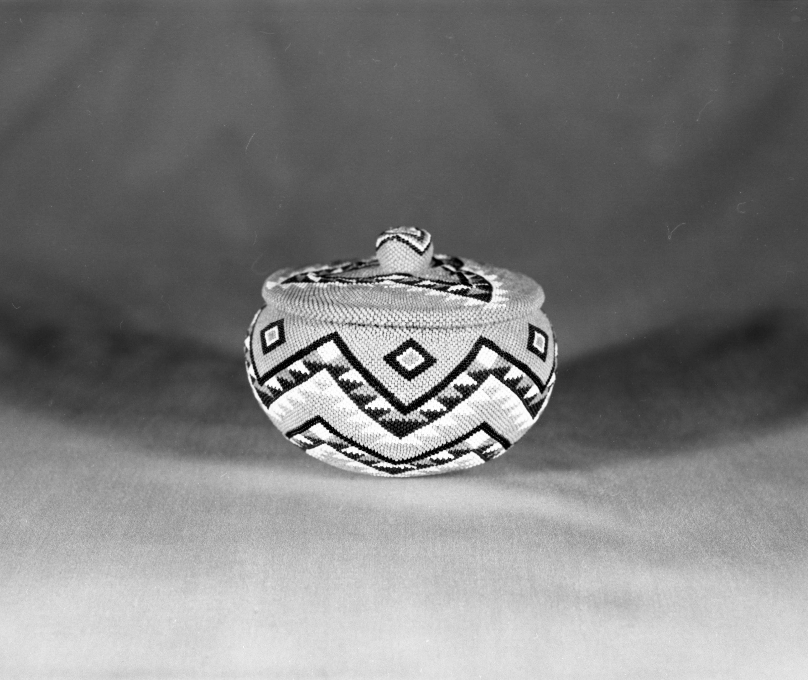 Private Collection. Object: Paiute basket. Dimensions: 5 1/4 x 4". Materials: Willow, glass beads (probably Czech), cotton thread. Pale green background, dark blue, medium blue, red, yellow & yellow-orange. Technology: Non-interlocking stitch, fag ends trimmed on interior, rim of main basket overstitched twice. Collection History: Made by Carrie Bethel, purchased from her by Donald Segerstrom, c. 1960. Photographed by Michael Dixon. Catalogued by Craig Bates and Barbara Beroza. Yosemite Collections, National Park Service.
