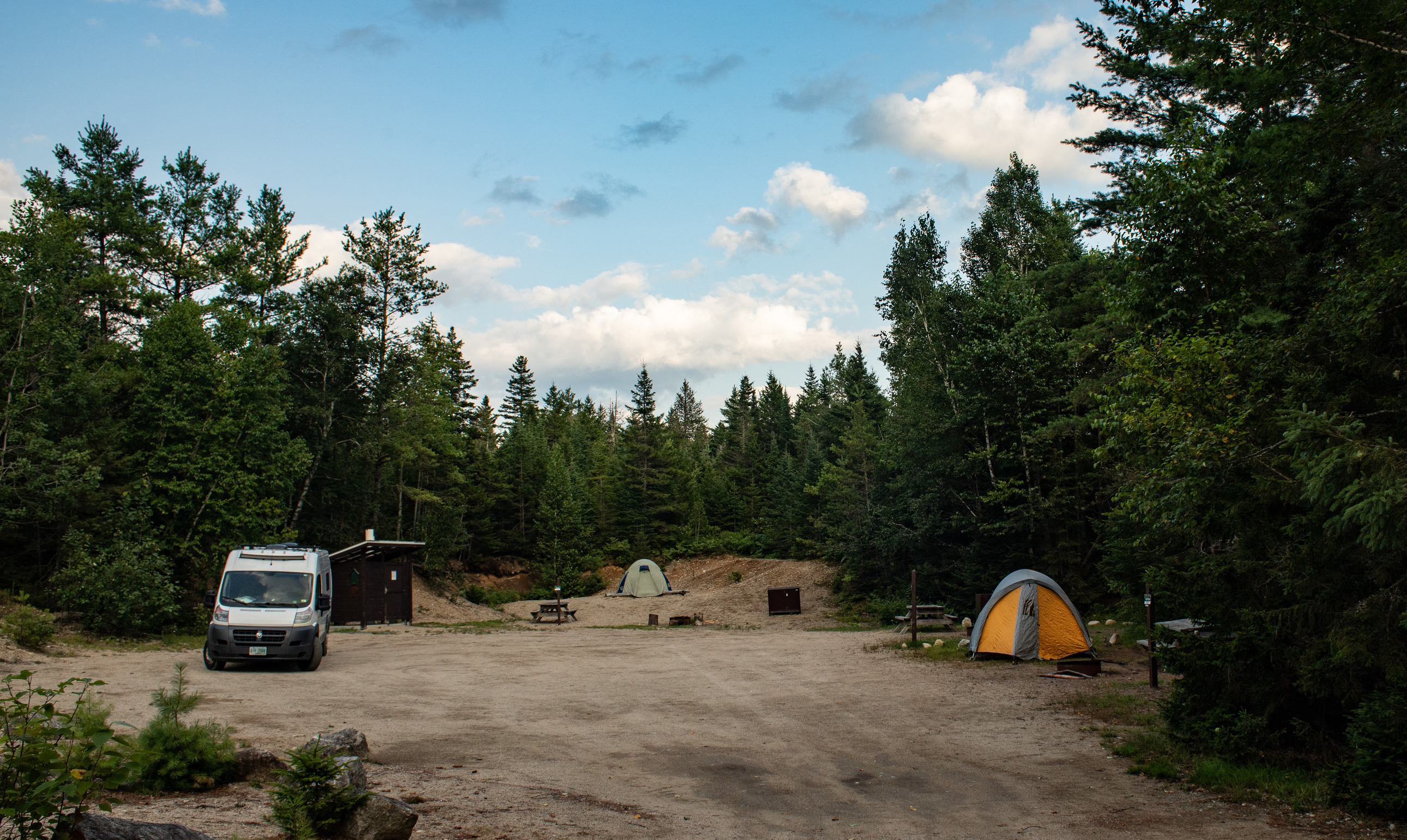 Two tents and a van are set up along the sides of a sandy campground surrounded by pine trees