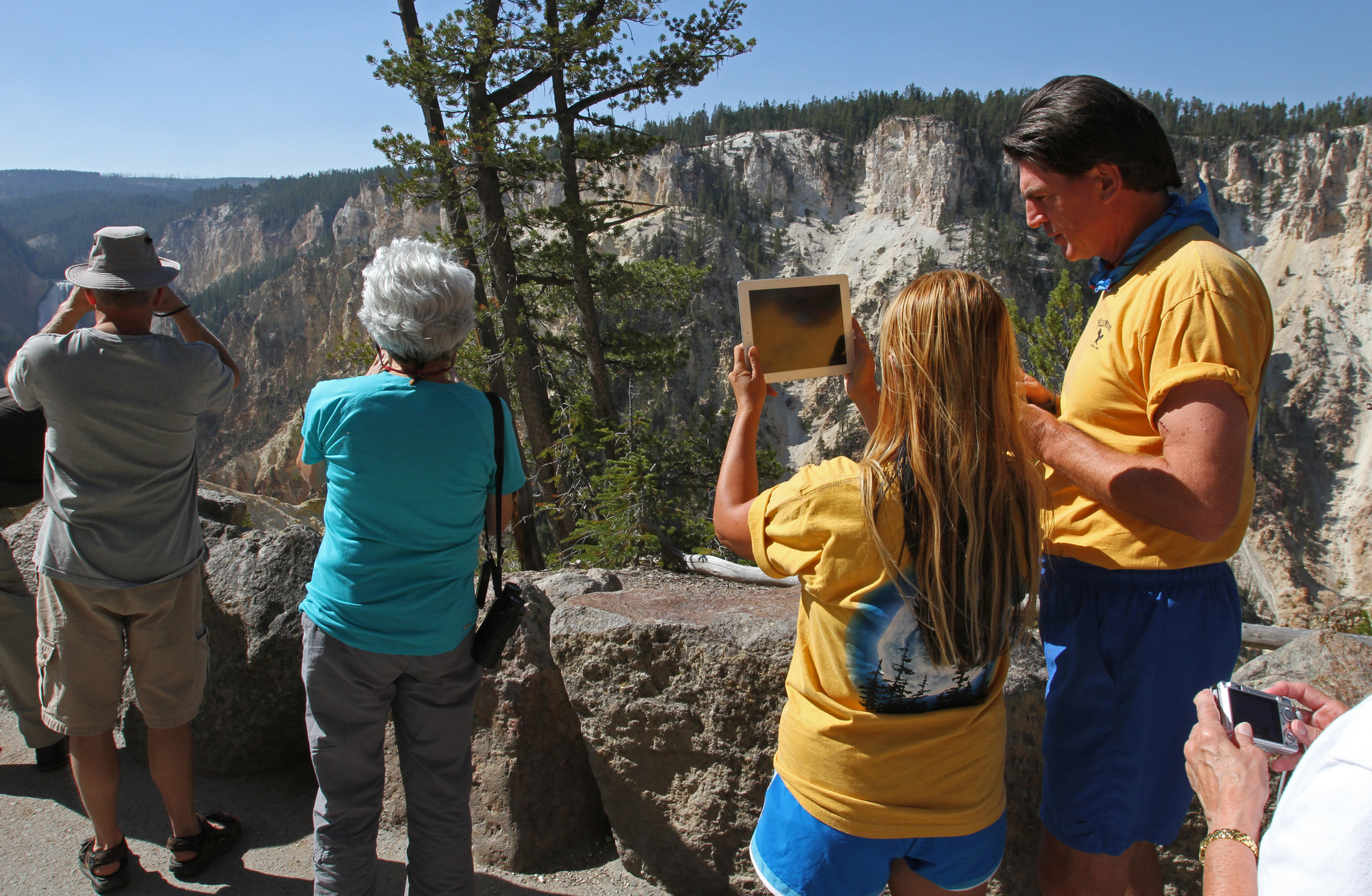 A man and a woman at an overlook.  The woman is holding up an ipad in the direction of the Lower Falls which are not in view.