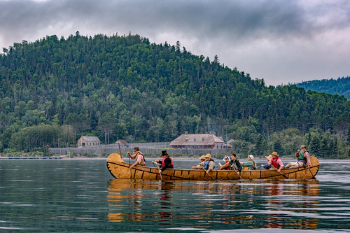 A group of people paddle a Montreal Canoe in Grand Portage Bay. In the distance a historic fur trade post.