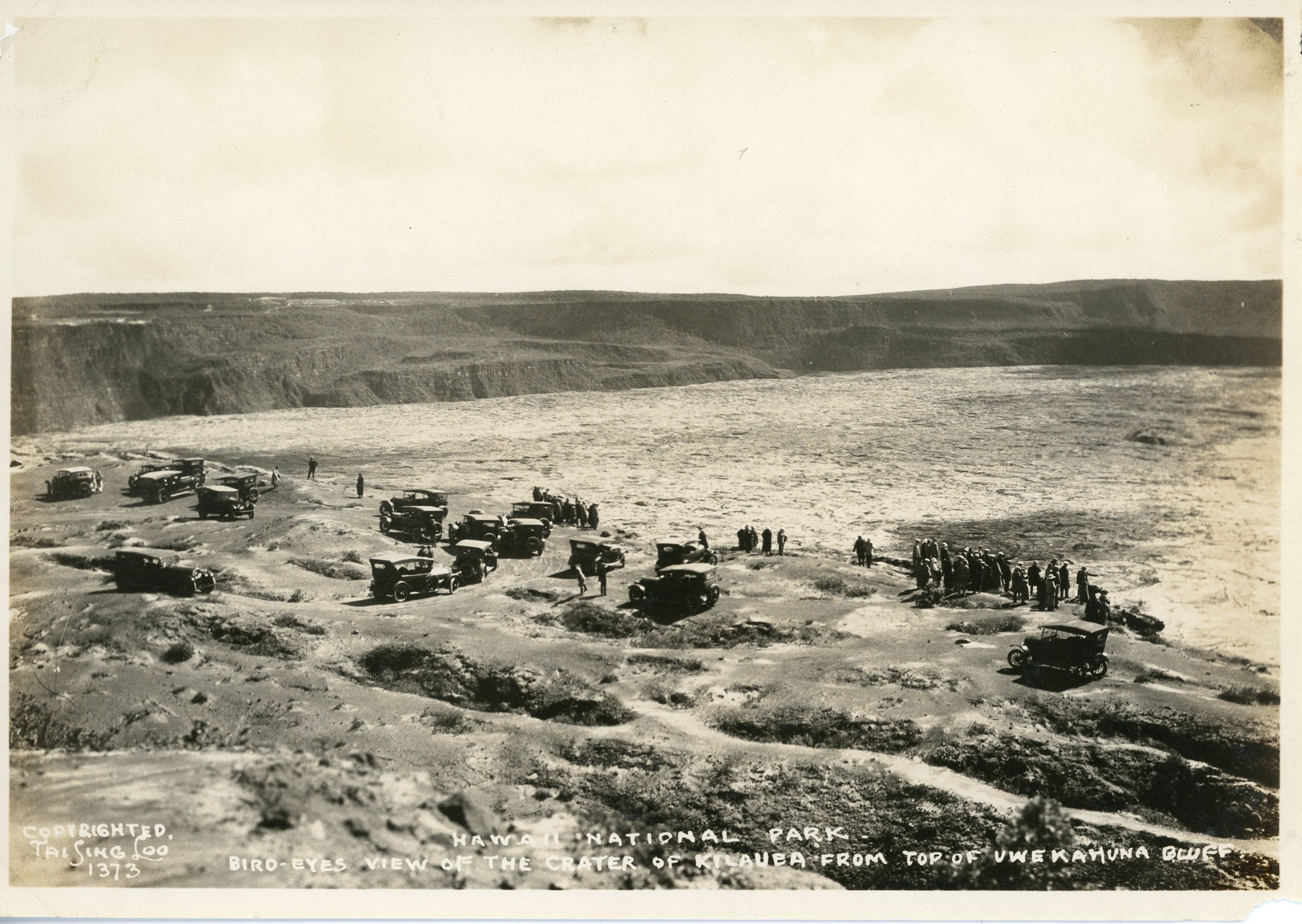 A black and white image of Kīlauea from Uwēkahuna bluff. The image was taken from afar and shows many cars parked along the caldera floor. There are eighteen cars parked sporadically around the area with some visitors standing near the cars while others are standing along the edge of a crater looking at the lava lake. In the distance the walls of the caldera are visible.  On the left bottom corner of the image there is a photographer signature, “Copyrighted Tai Sing Loo 1373.” Also along the bottom of the image is a caption that reads, “Hawaii National Park. Bird’s-eye view of the crater of Kilauea from top of Uwekahuna Bluff.”